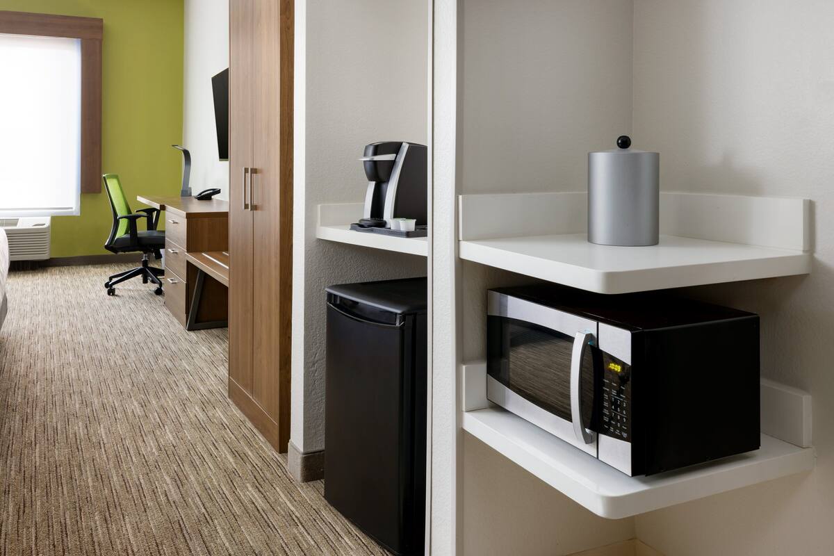 A hotel room with a compact kitchenette, a black refrigerator, a microwave, a coffee maker, and a green office chair.