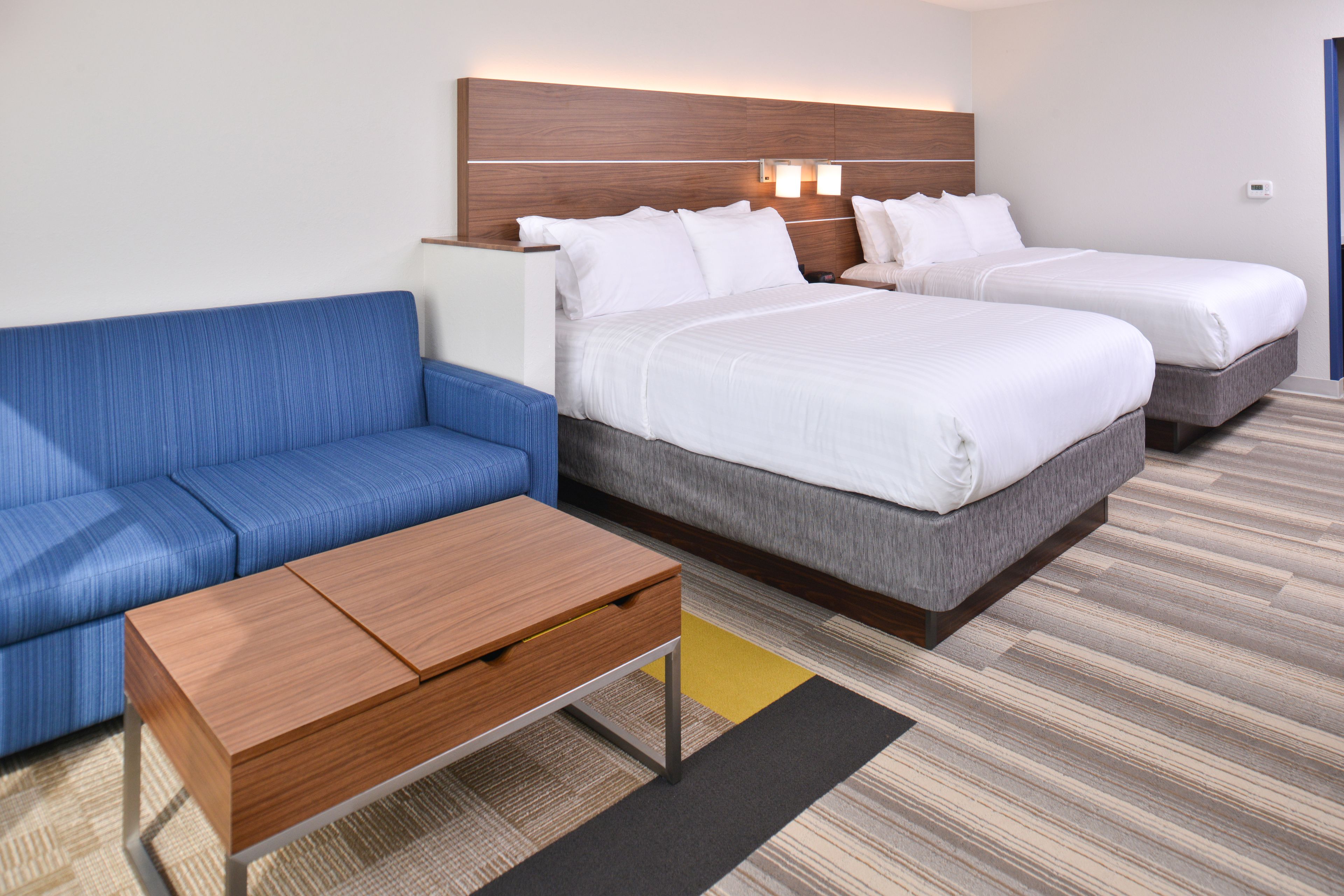 Une chambre d’hôtel avec un canapé bleu, une table en bois, deux lits avec du linge de lit blanc et une tête de lit dotée d’un éclairage intégré.