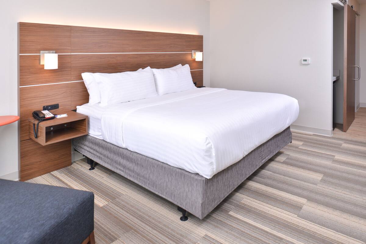 A modern hotel room with a large bed, a wooden headboard, bedside lamps, a nightstand, and a grey upholstered bench.