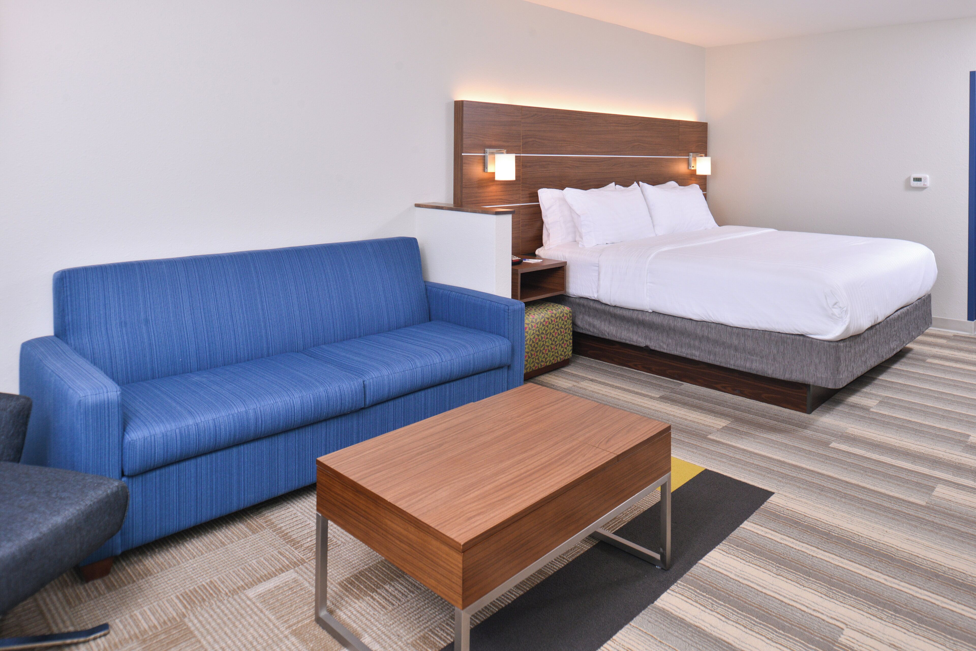 A hotel room with a blue sofa, a wooden coffee table, a bed with white bedding, and a nightstand with a lamp.