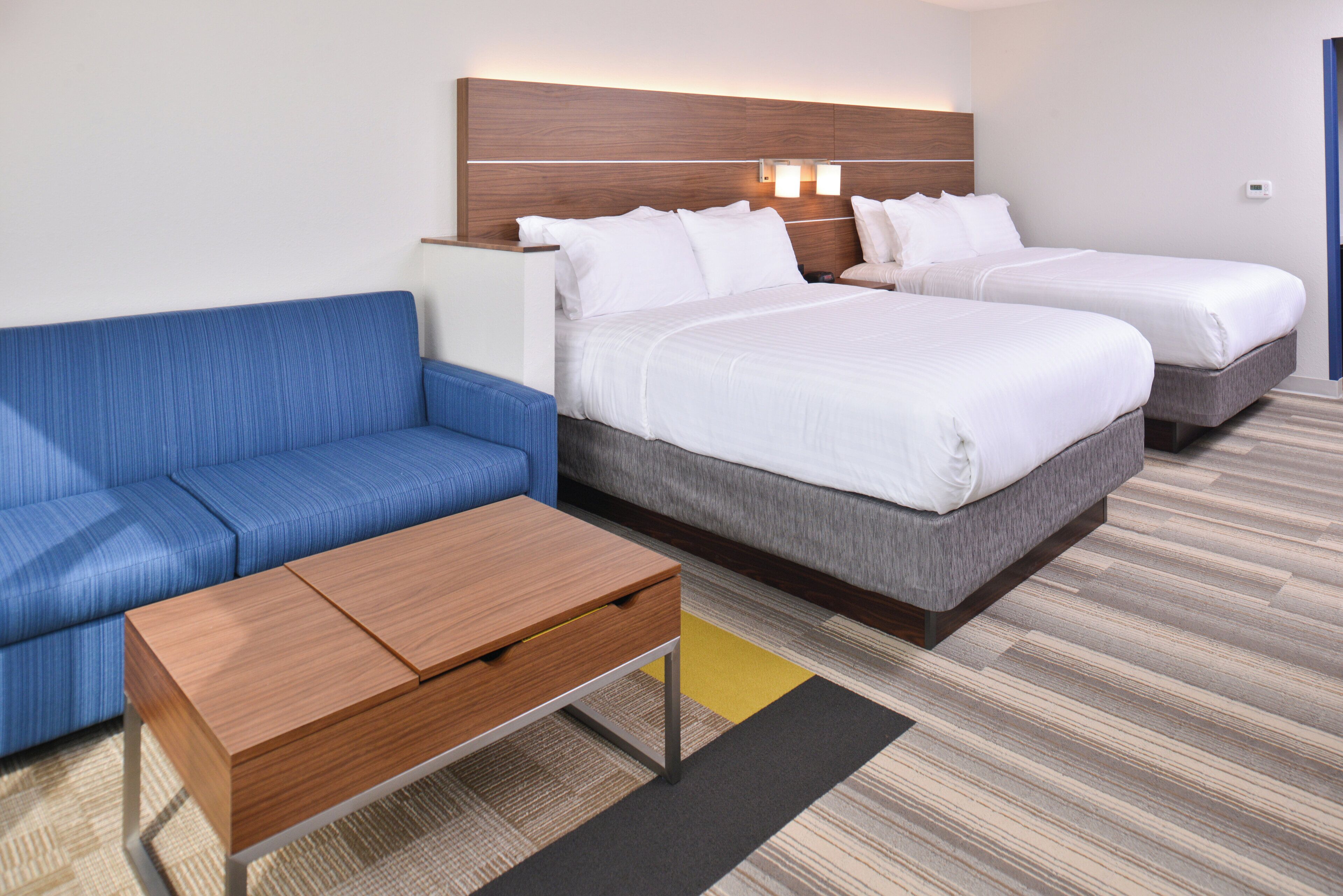 A hotel room with a blue sofa, two single beds, a wooden nightstand, and a small table.