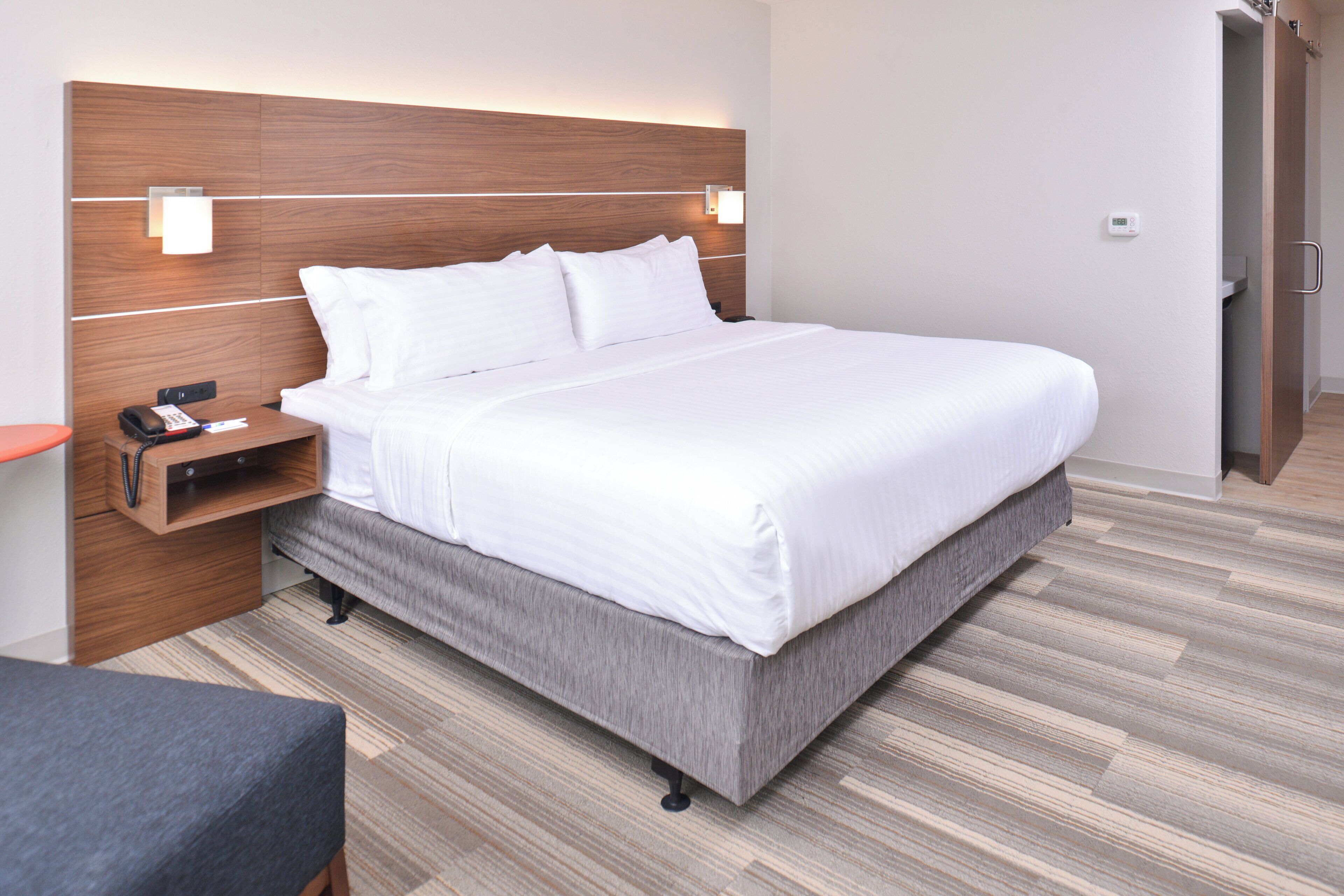 Quarto de hotel moderno com uma cama grande, uma mesinha de cabeceira com telefone e papéis, um criado-mudo e um guarda-roupa.