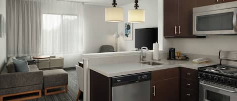 A compact hotel kitchen with a dishwasher, stove, microwave, and a small dining area with a sofa and table.