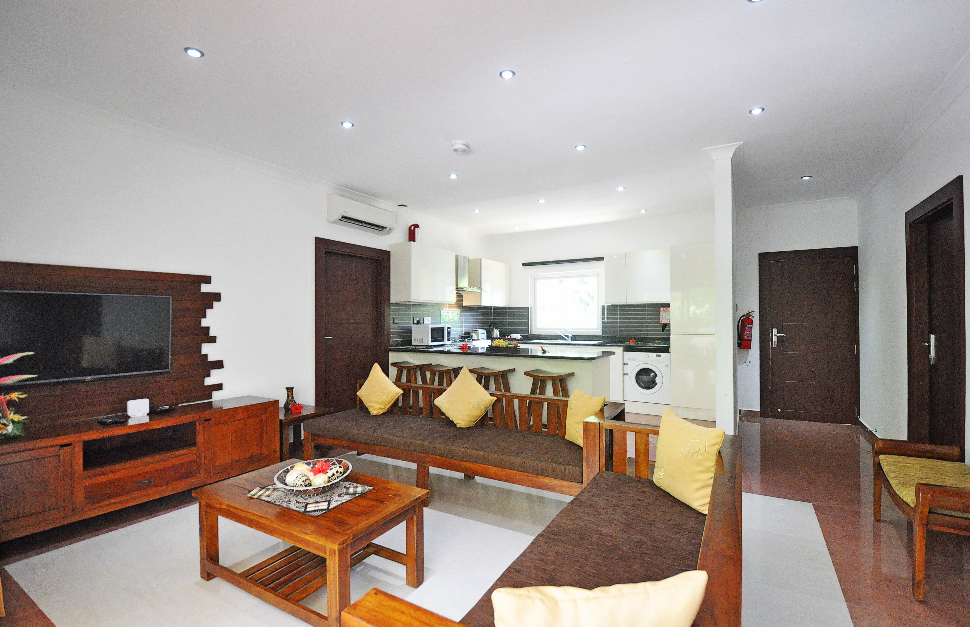 A modern living room with a flat-screen TV, wooden furniture, and a kitchen in the background.