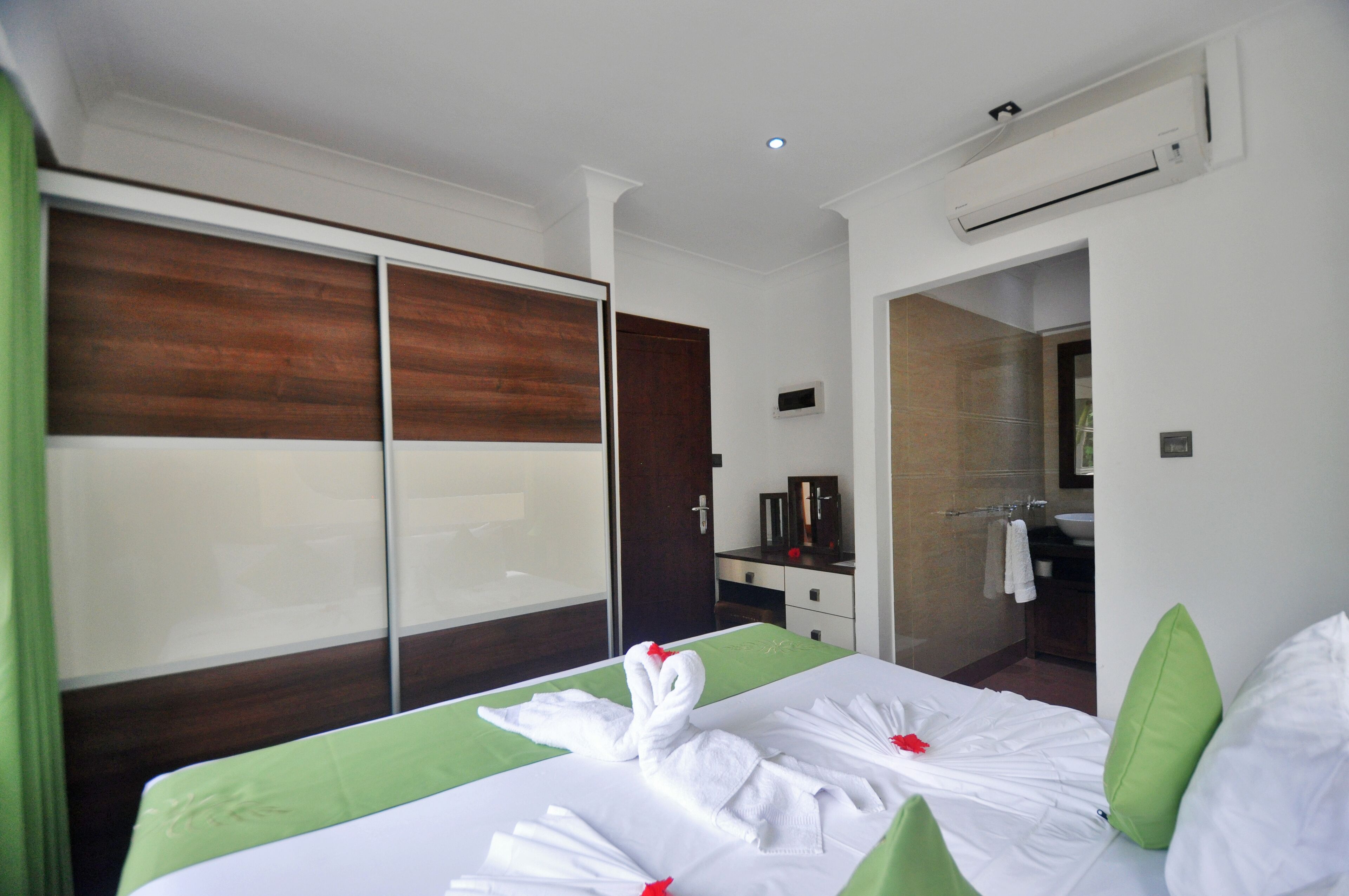 A hotel room with a bed, a green bedspread, a wooden headboard, a bathroom with a sink and mirror, and an air conditioning unit.