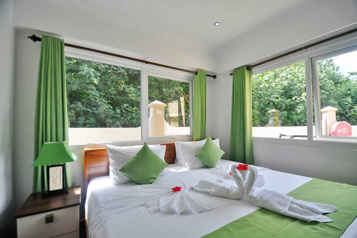 Ein Hotelzimmer mit einem ordentlich bezogenen Bett, grünen Vorhängen und Blick ins Grüne.