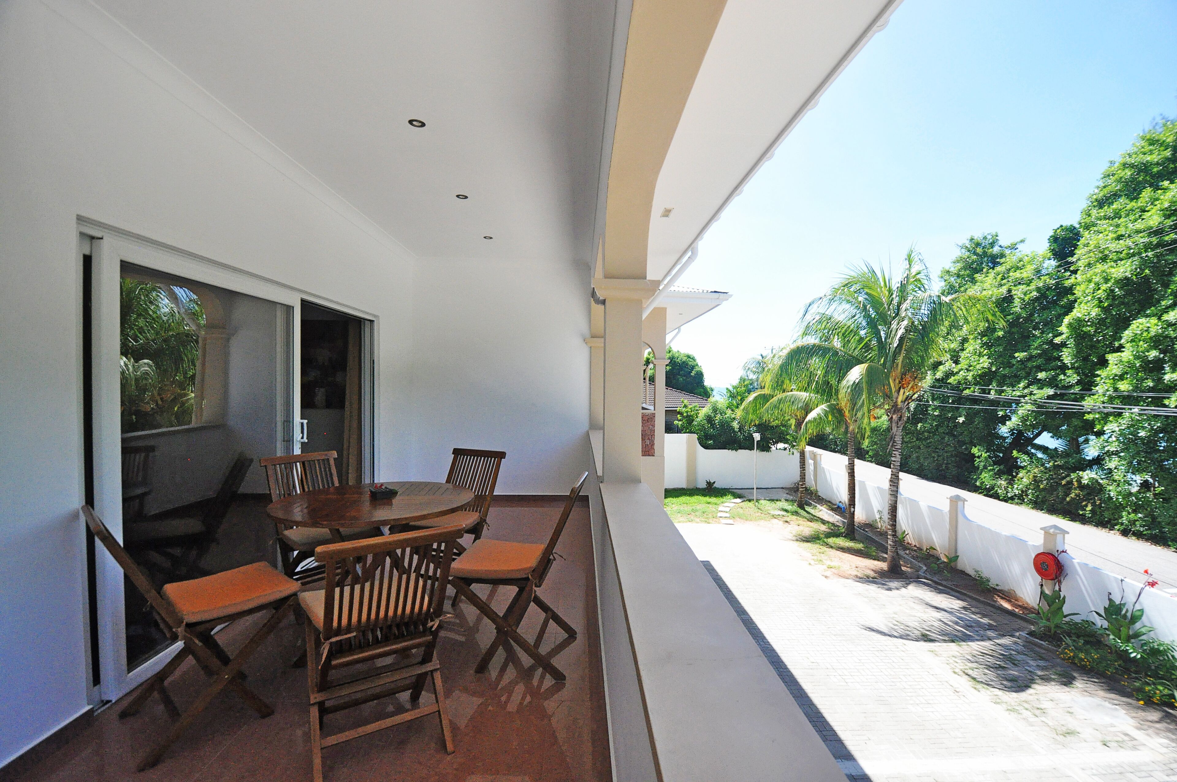 A balcony with a wooden table and chairs, a sliding glass door, and a view of a white wall and greenery.