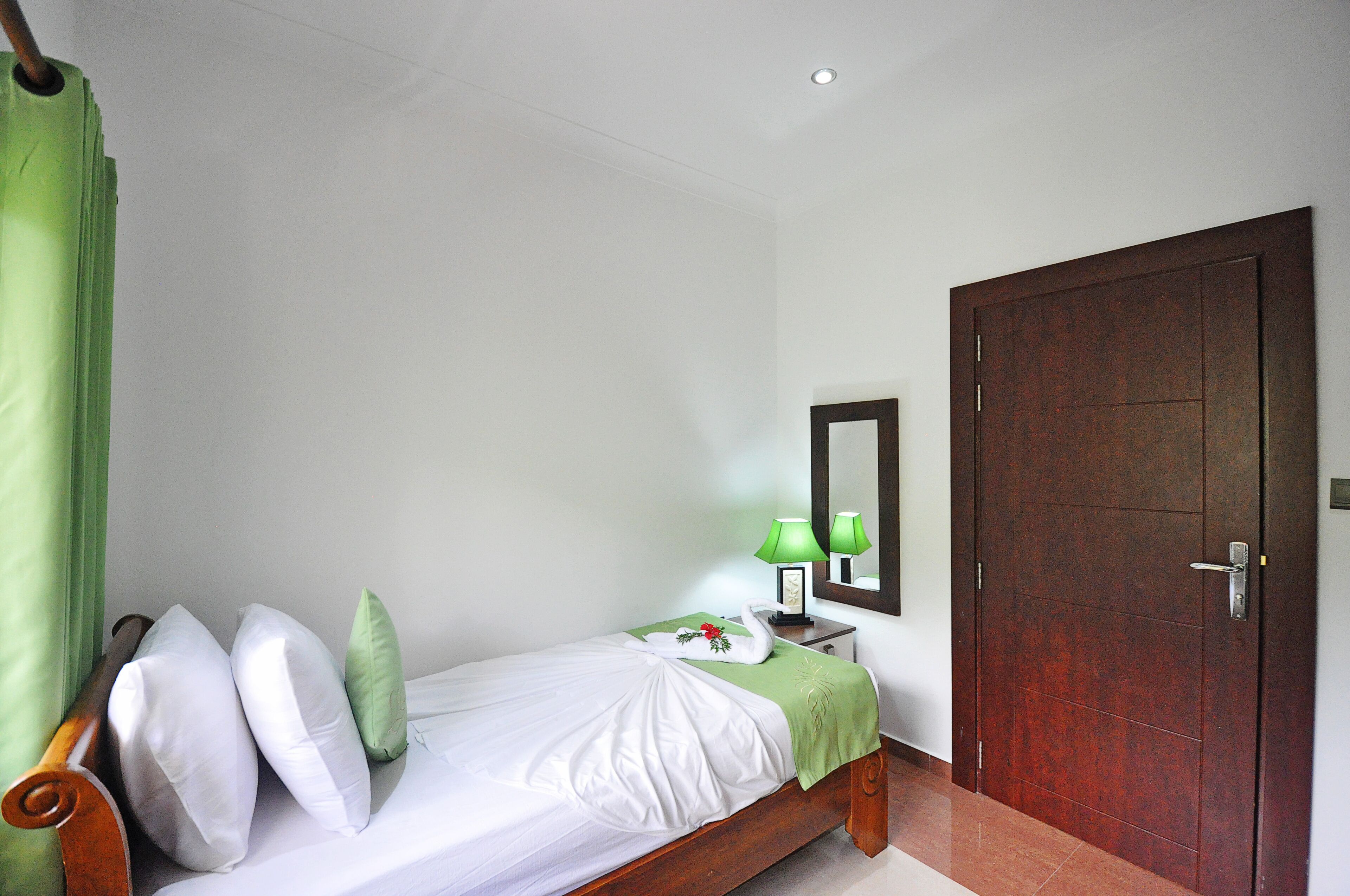 A neatly made bed with pillows, a green bedspread, and a swan decoration. There is a nightstand with a lamp and a framed picture. A wooden door and a mirror are on the wall.