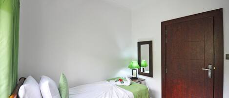 A neatly made bed with pillows, a green bedspread, and a swan decoration. There is a nightstand with a lamp and a framed picture. A wooden door and a mirror are on the wall.