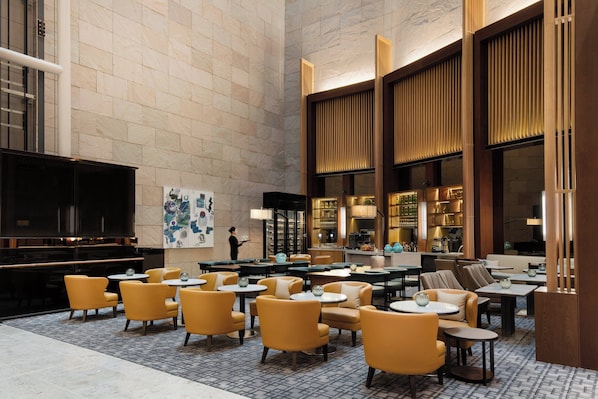 A spacious lobby with a high ceiling, marble walls, and a mix of seating areas including yellow armchairs and tables.