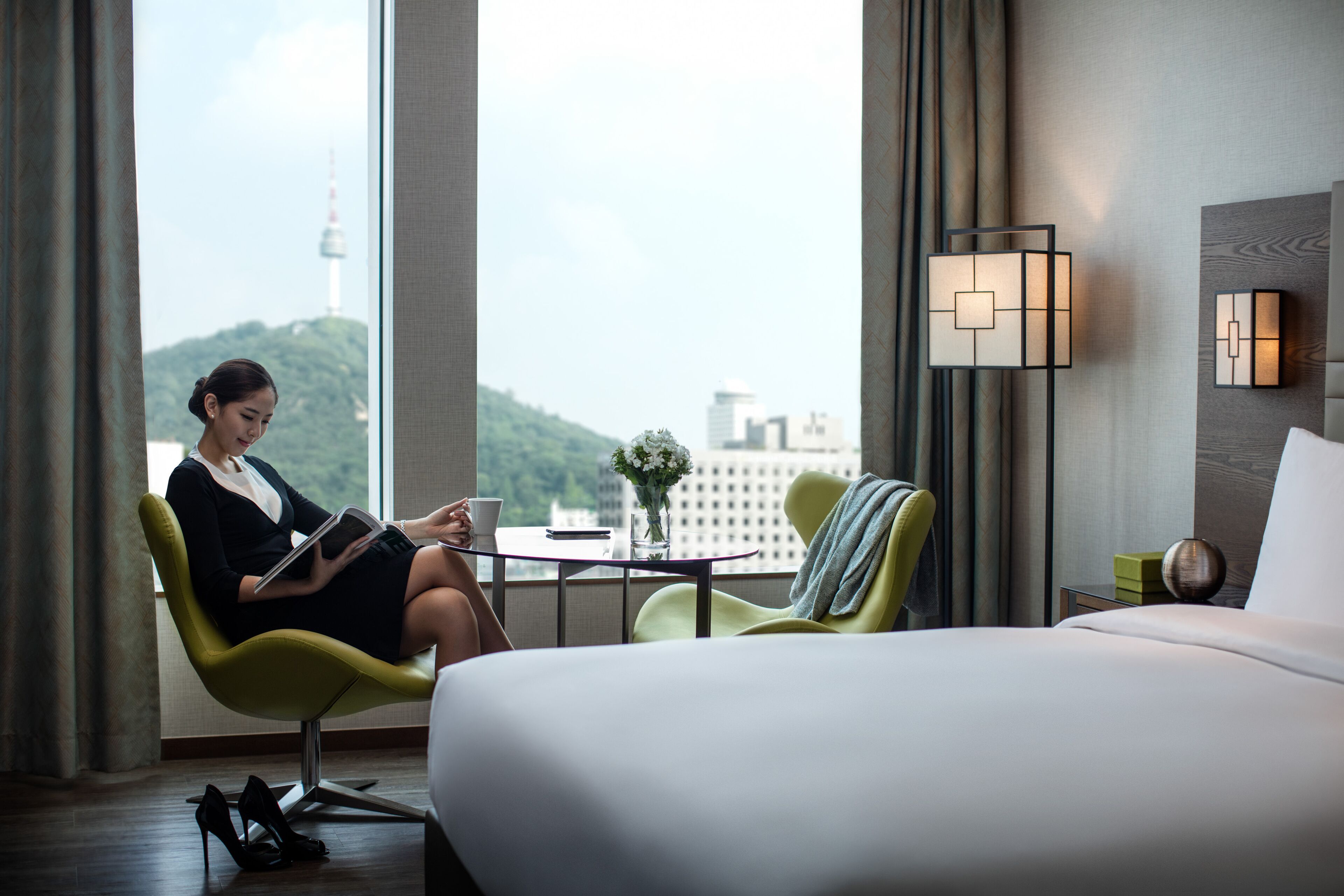 A hotel room with a bed, a green armchair, a small table with a vase of flowers, and a view of a cityscape with a tower.