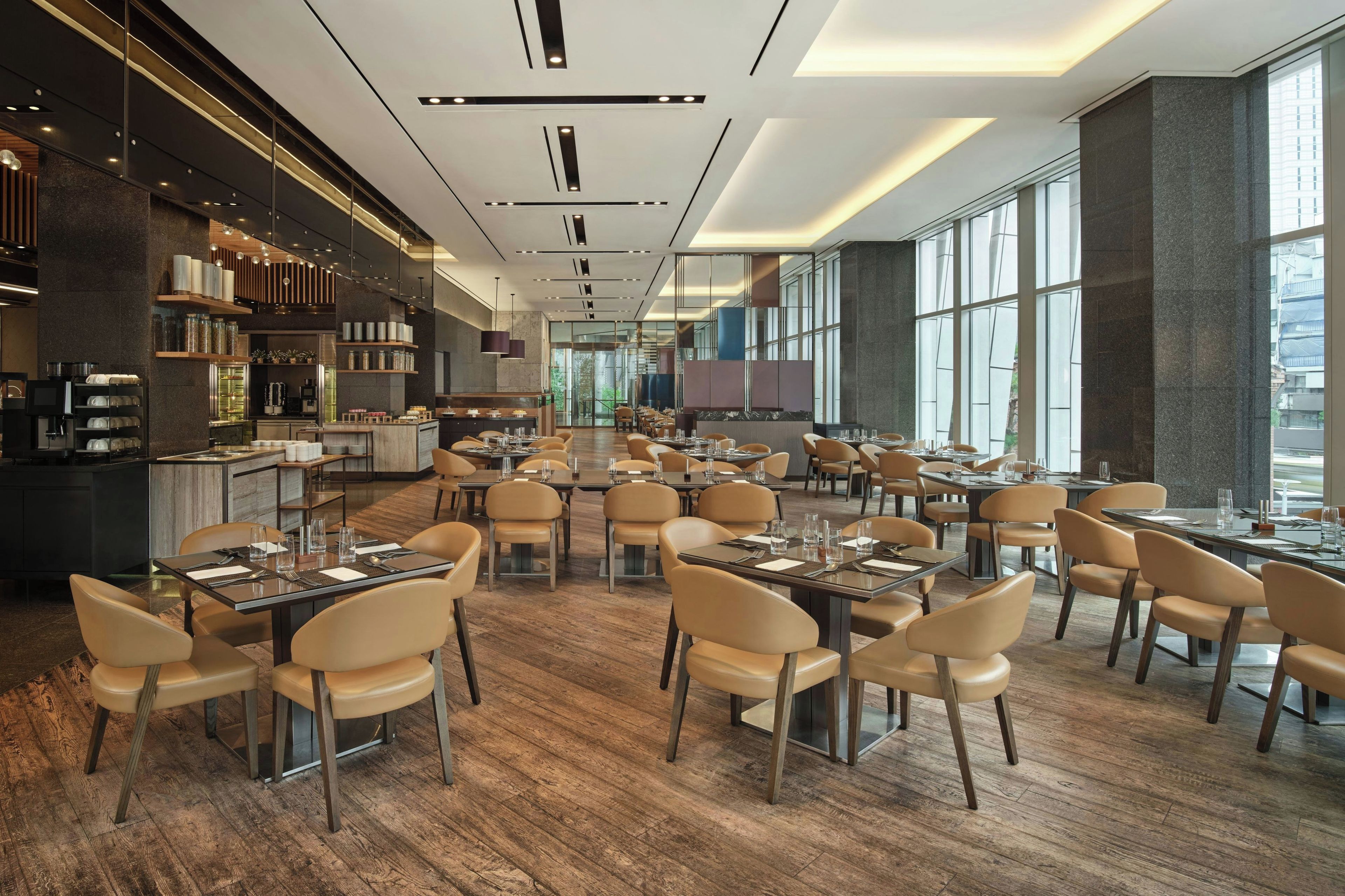 A modern restaurant with multiple dining tables and chairs, a kitchen area, and large windows providing natural light.