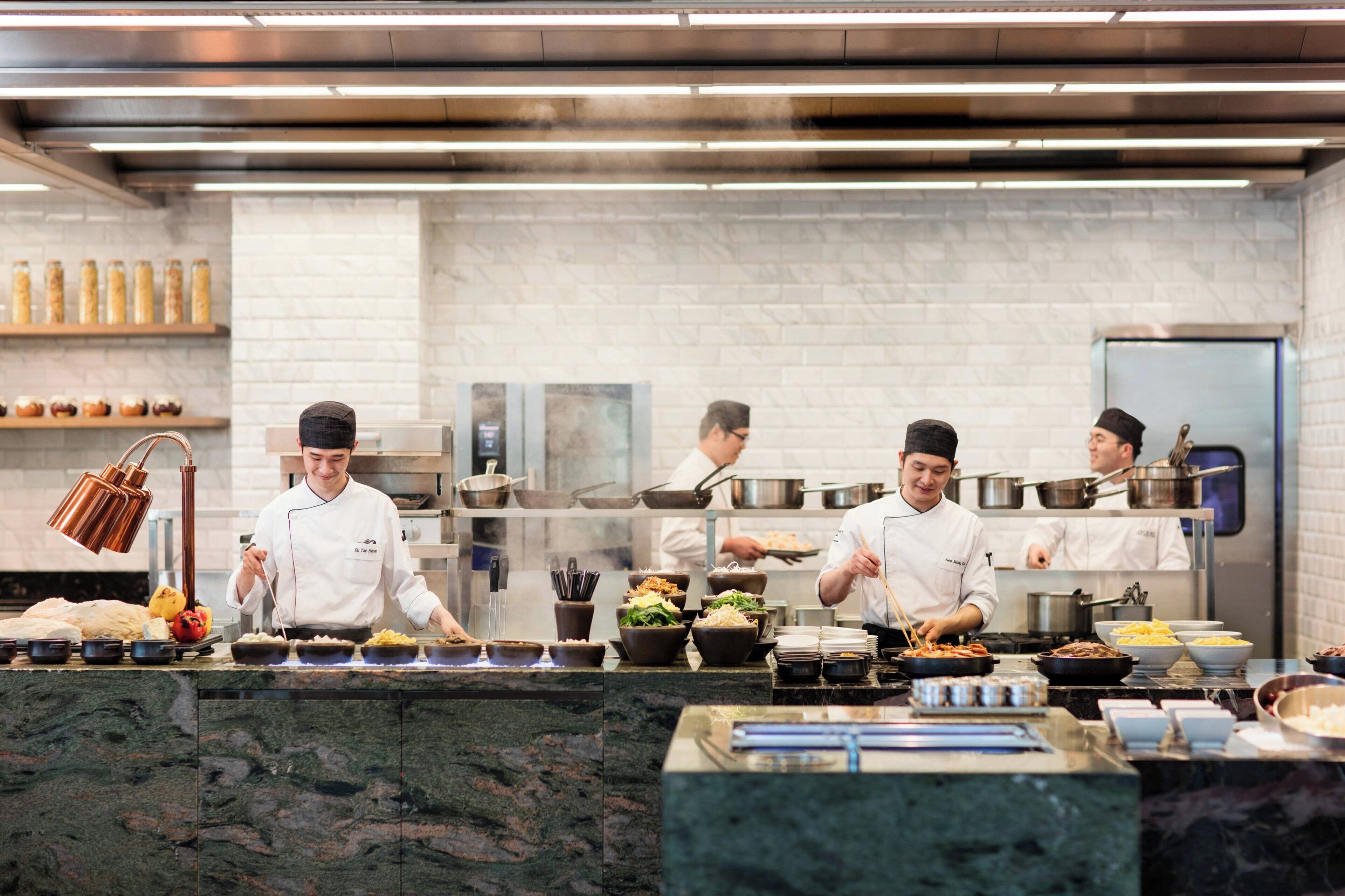 A professional kitchen with chefs preparing food, stainless steel appliances, and a marble countertop.