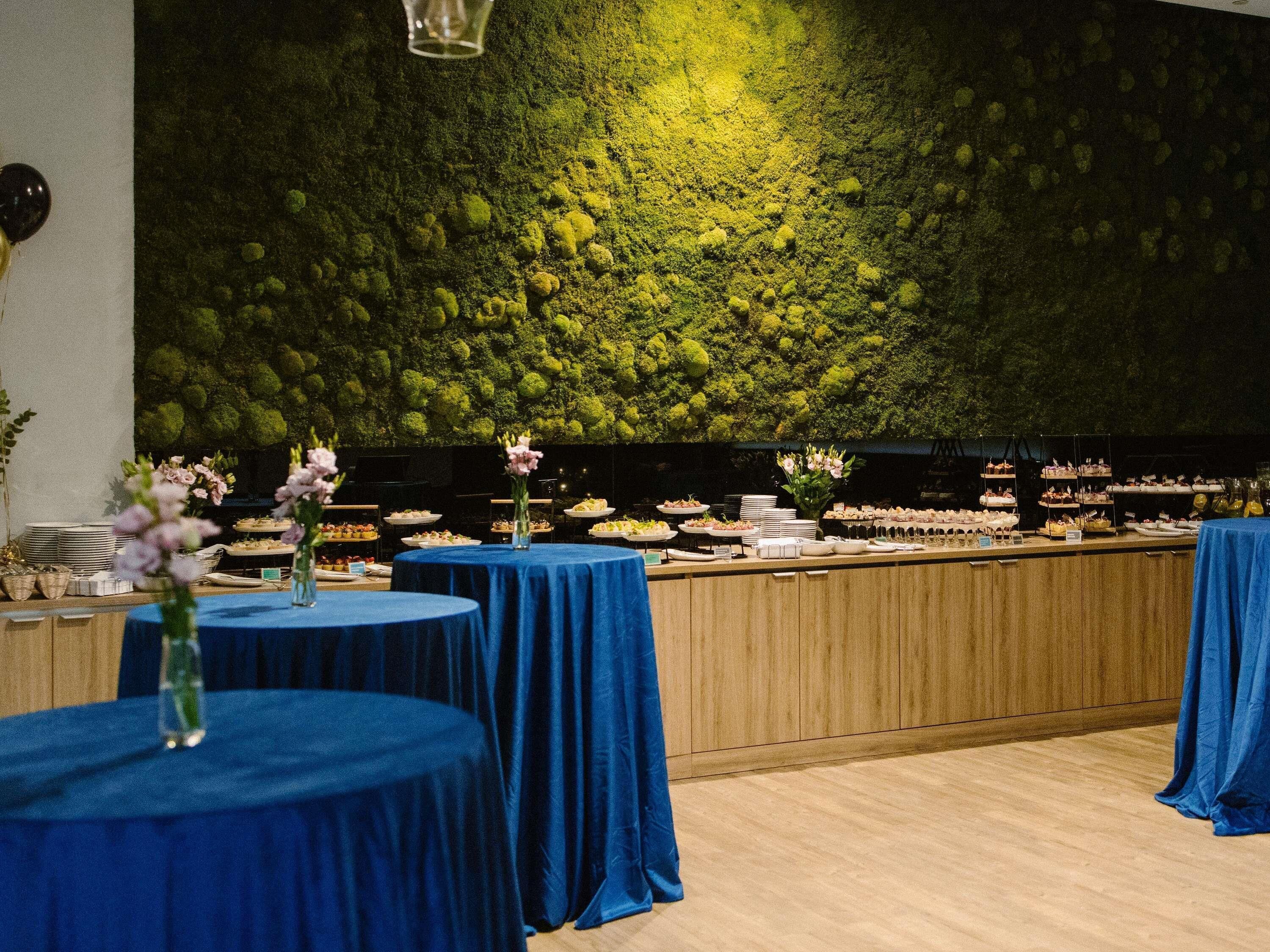 Un restaurant avec des tables rondes recouvertes de nappes bleues, un mur végétal vert et différents plats exposés sur des étagères.