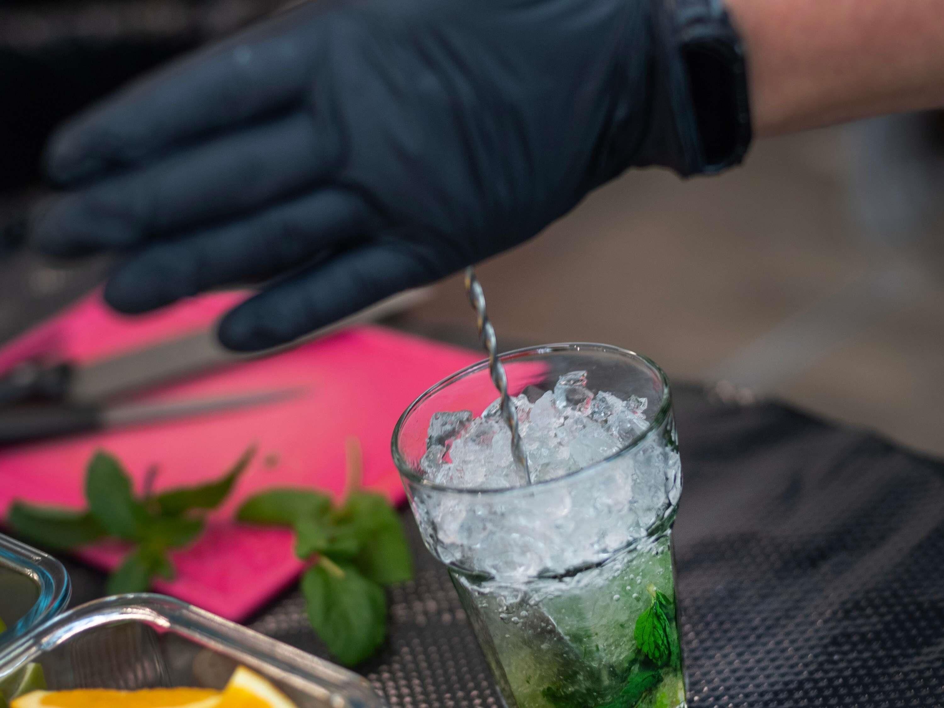 A person wearing a black glove pouring a liquid into a glass with ice and mint leaves.