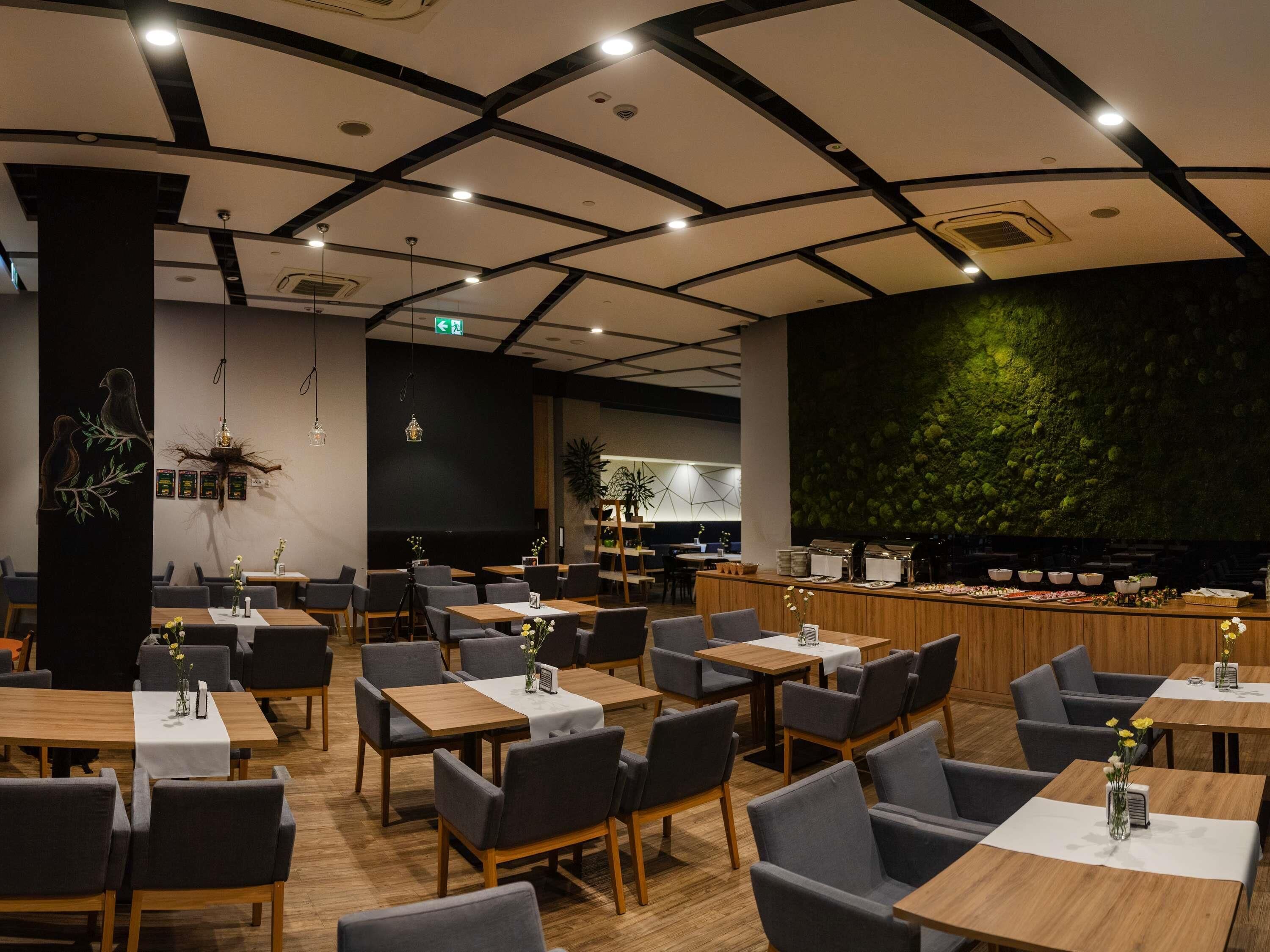 A modern restaurant with a high ceiling, wooden tables, and grey chairs. There is a green wall with a textured pattern and a bar area with a wooden counter.