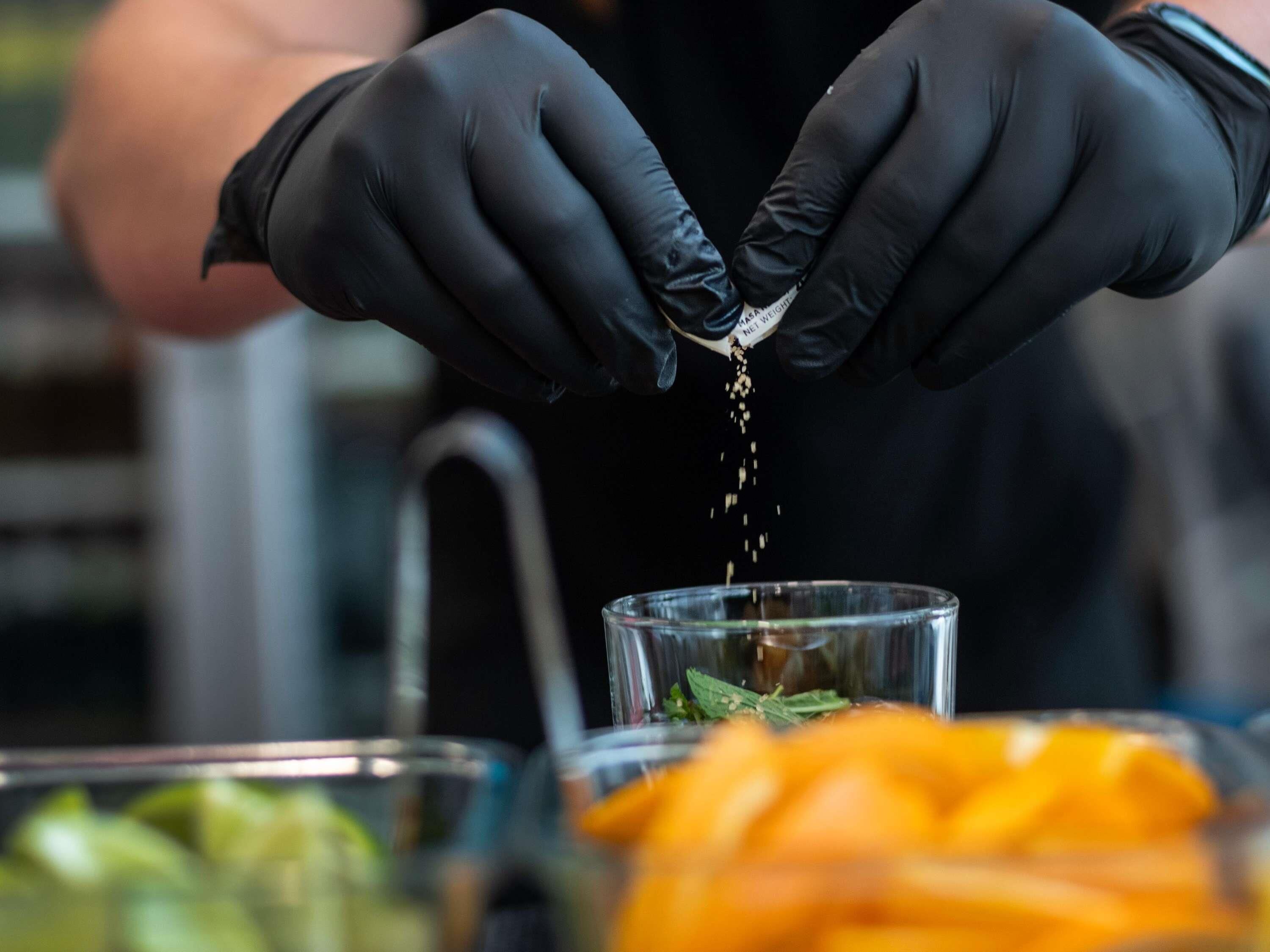 Hands wearing black gloves sprinkling salt over a dish with herbs and vegetables.