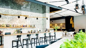 Zona de bar moderna con pared de diseño geométrico, una barra alargada con taburetes y una variedad de botellas y copas.