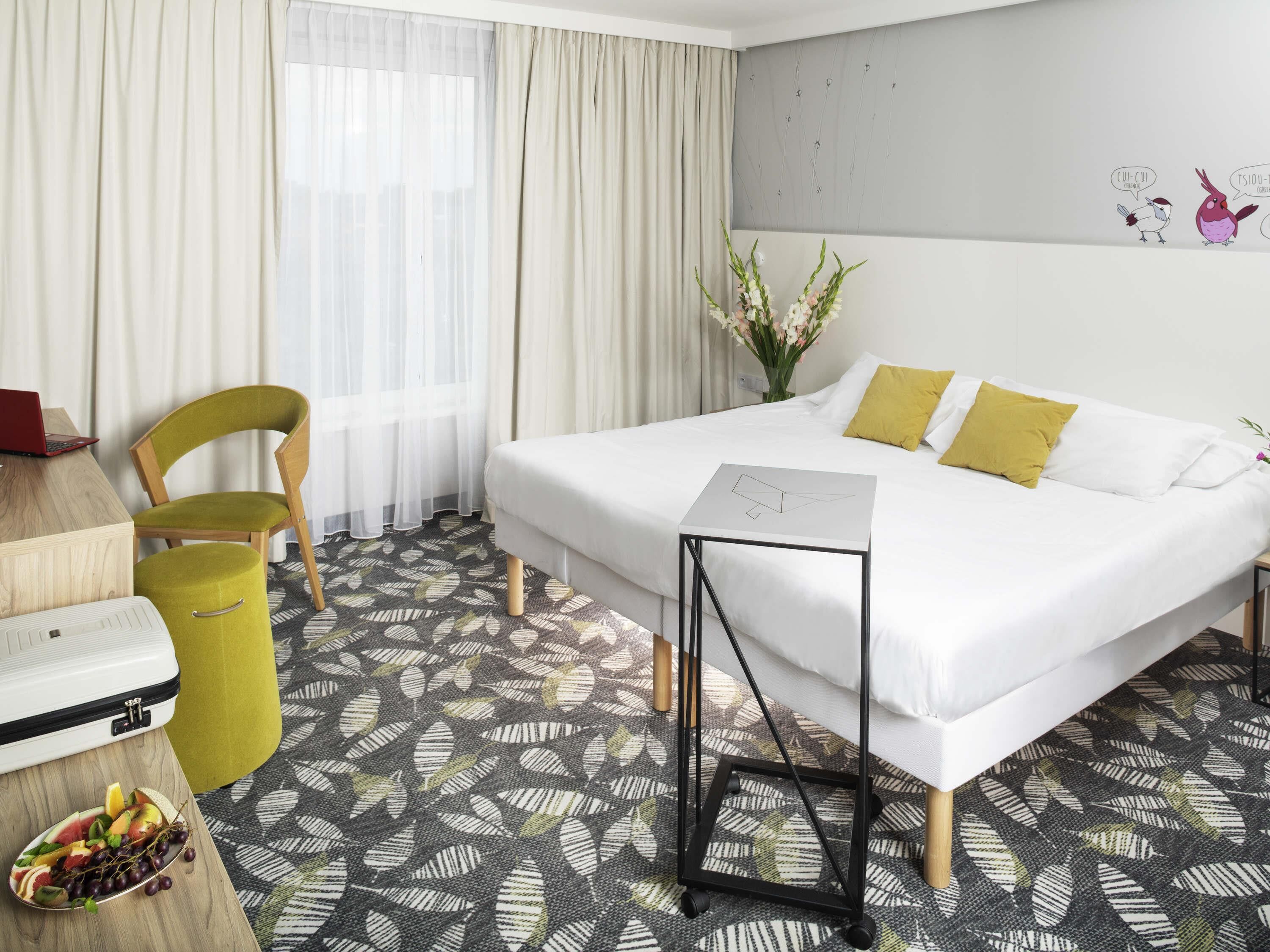 A hotel room with a bed, a desk with a laptop, a chair, a trash bin, and a fruit plate.