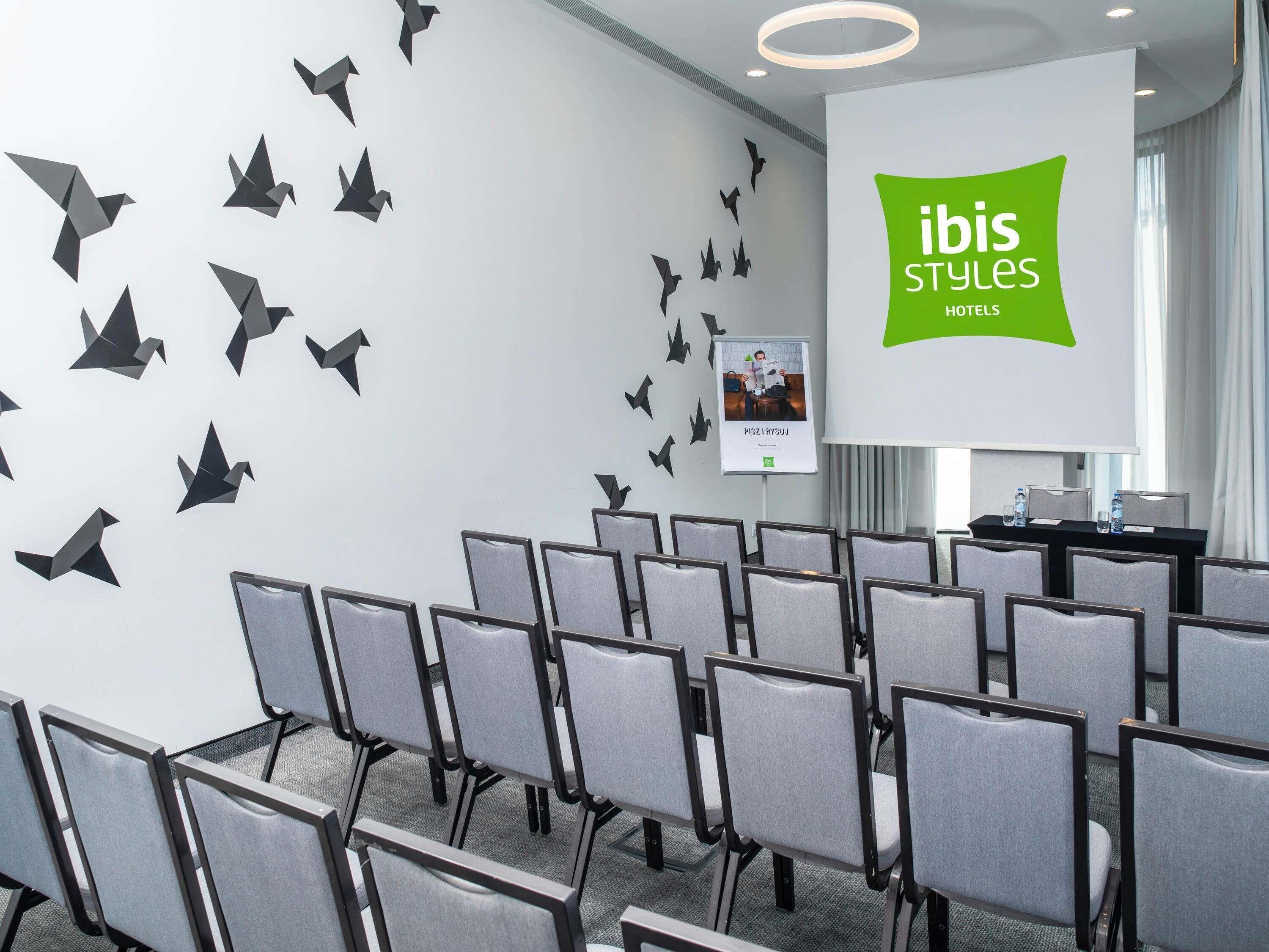 A conference room with rows of grey chairs, a whiteboard with 'ibis Styles Hotels' branding, and a projector screen.