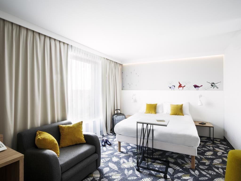 A modern hotel room with a bed, a sofa, a desk, and a window with curtains.