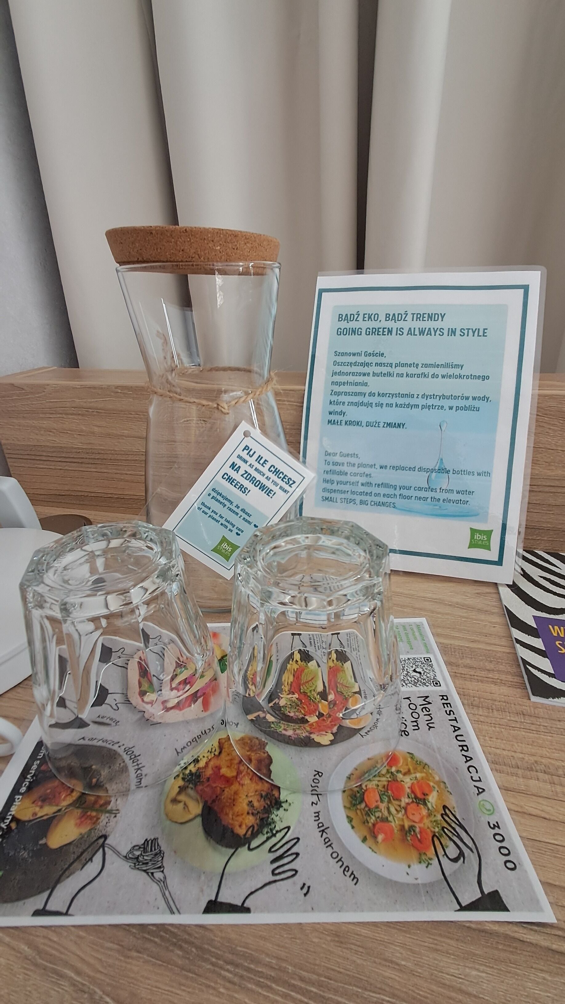A table with clear glasses, a cork stopper, a menu, and a sign promoting eco-friendly practices.