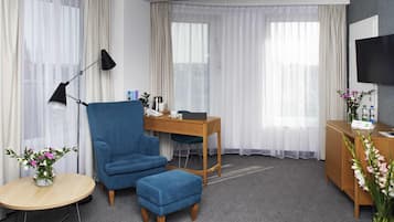 A hotel room with a blue armchair, a wooden desk, a small round table, a TV, and a bed.