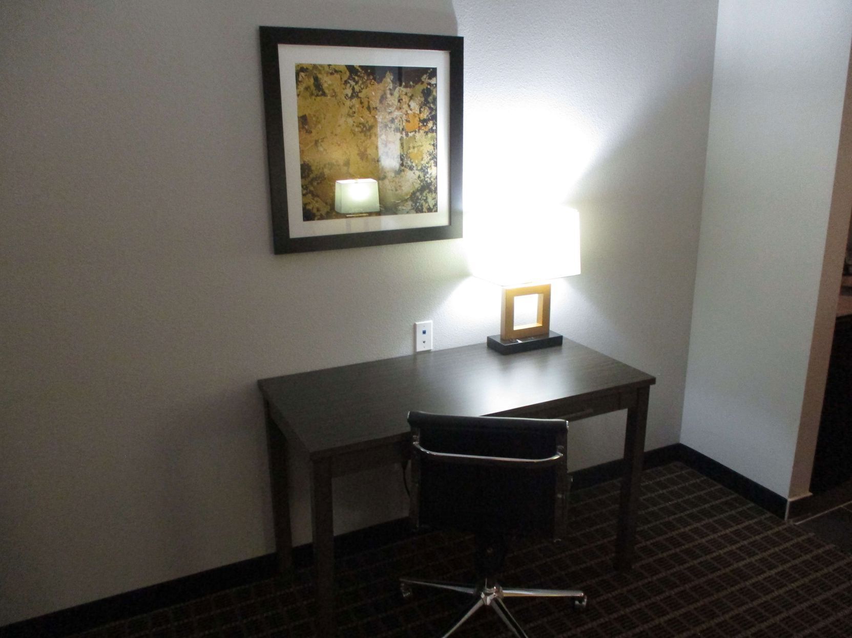 A hotel room with a desk, chair, framed artwork, and a lit lamp.