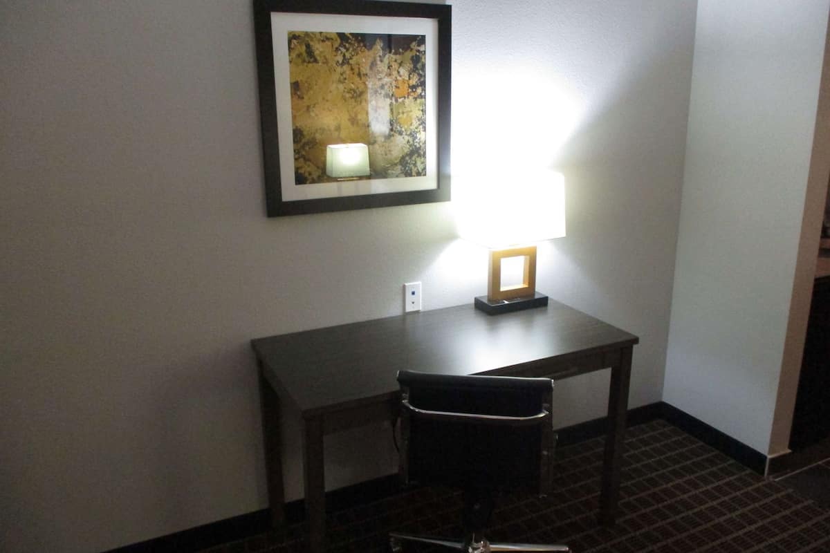 A hotel room with a desk, chair, framed artwork, and a lit lamp.