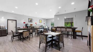 A modern dining area with multiple tables and chairs, a kitchen area with appliances, and a view of greenery outside.