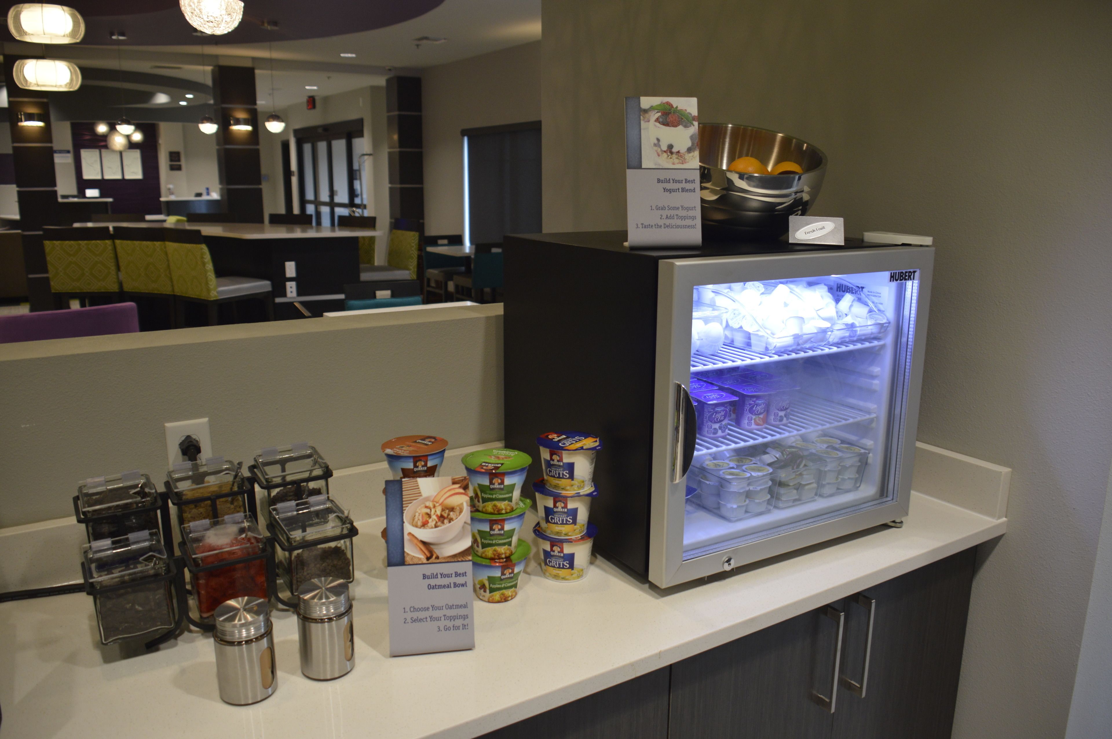 A self-service breakfast station with yogurt and cereal options, a refrigerator, and a coffee machine.