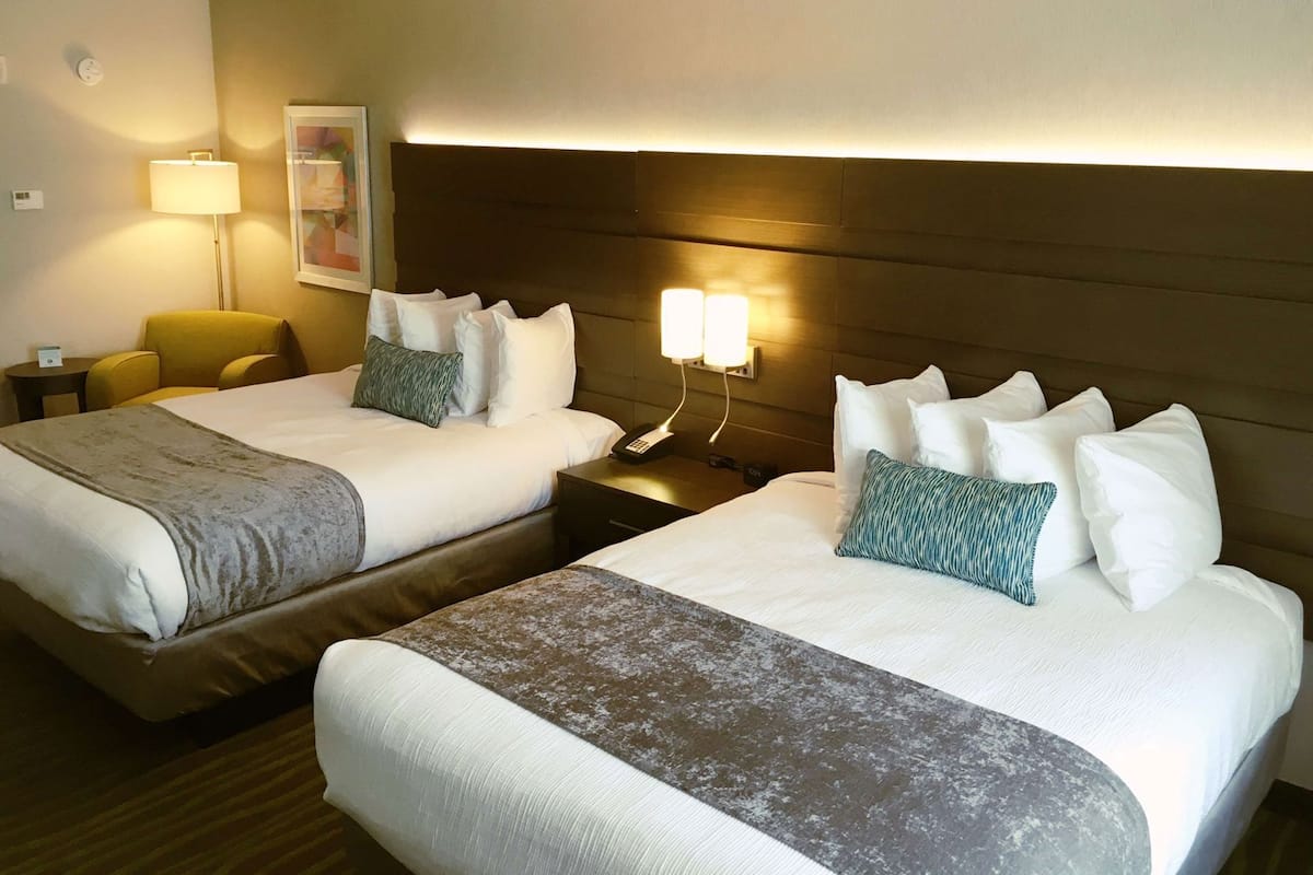 A hotel room with two beds, a bedside table with a lamp, a chair, and a wall-mounted light fixture.