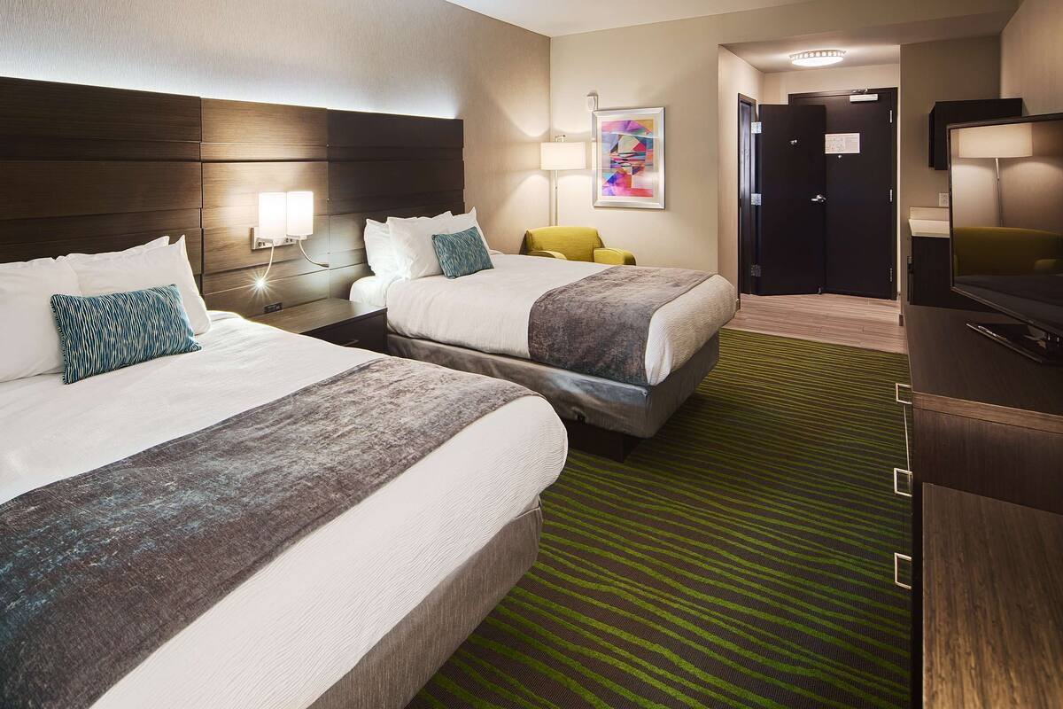 A hotel room with two beds, a green striped carpet, a wooden headboard, a nightstand with a lamp, and a painting on the wall.