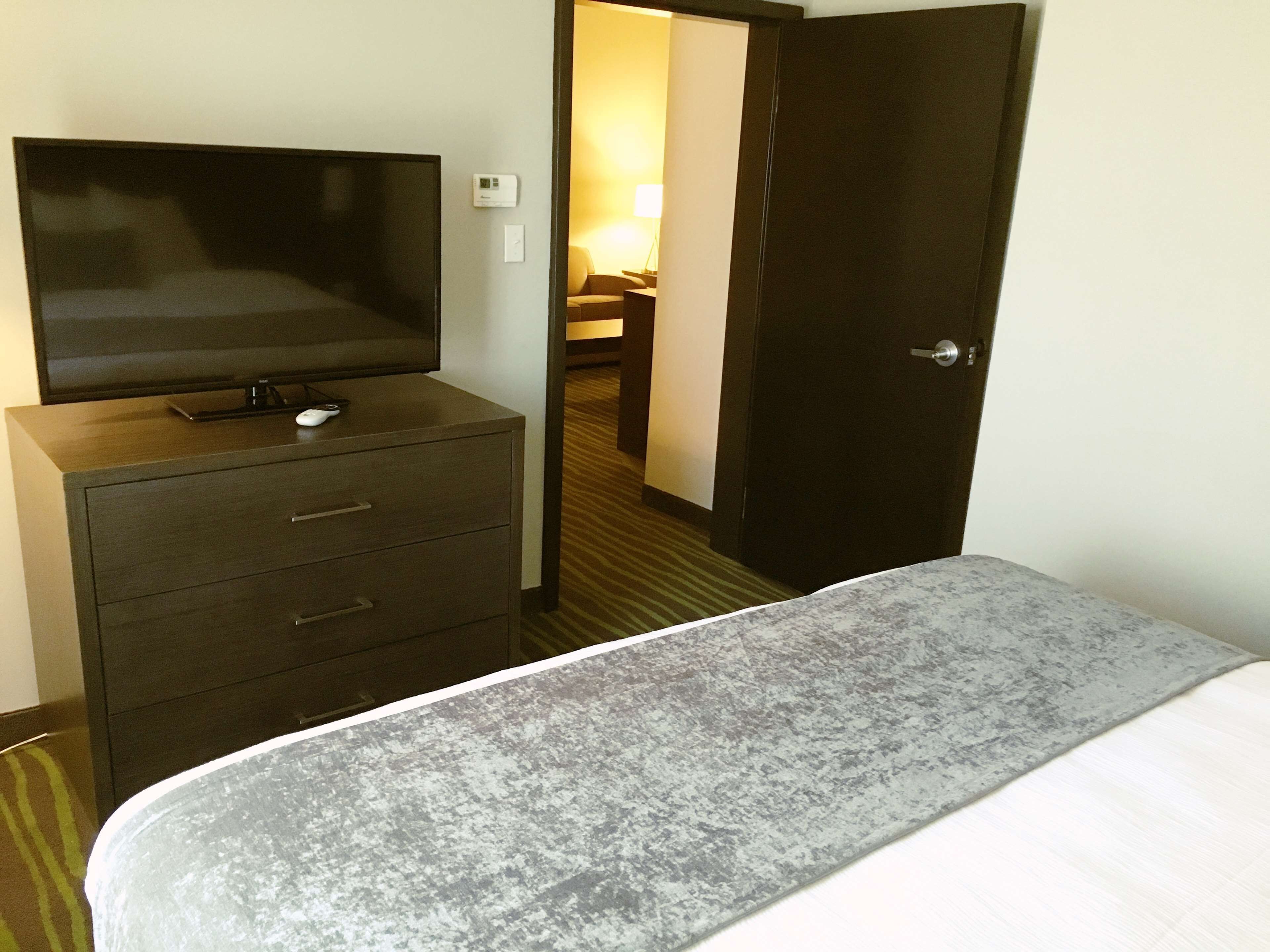 A hotel room with a bed, a TV on a dresser, a door, and a lamp in the background.