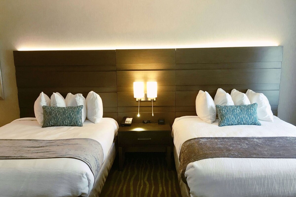A hotel room with two beds, a nightstand with a lamp, and a wall-mounted light fixture.