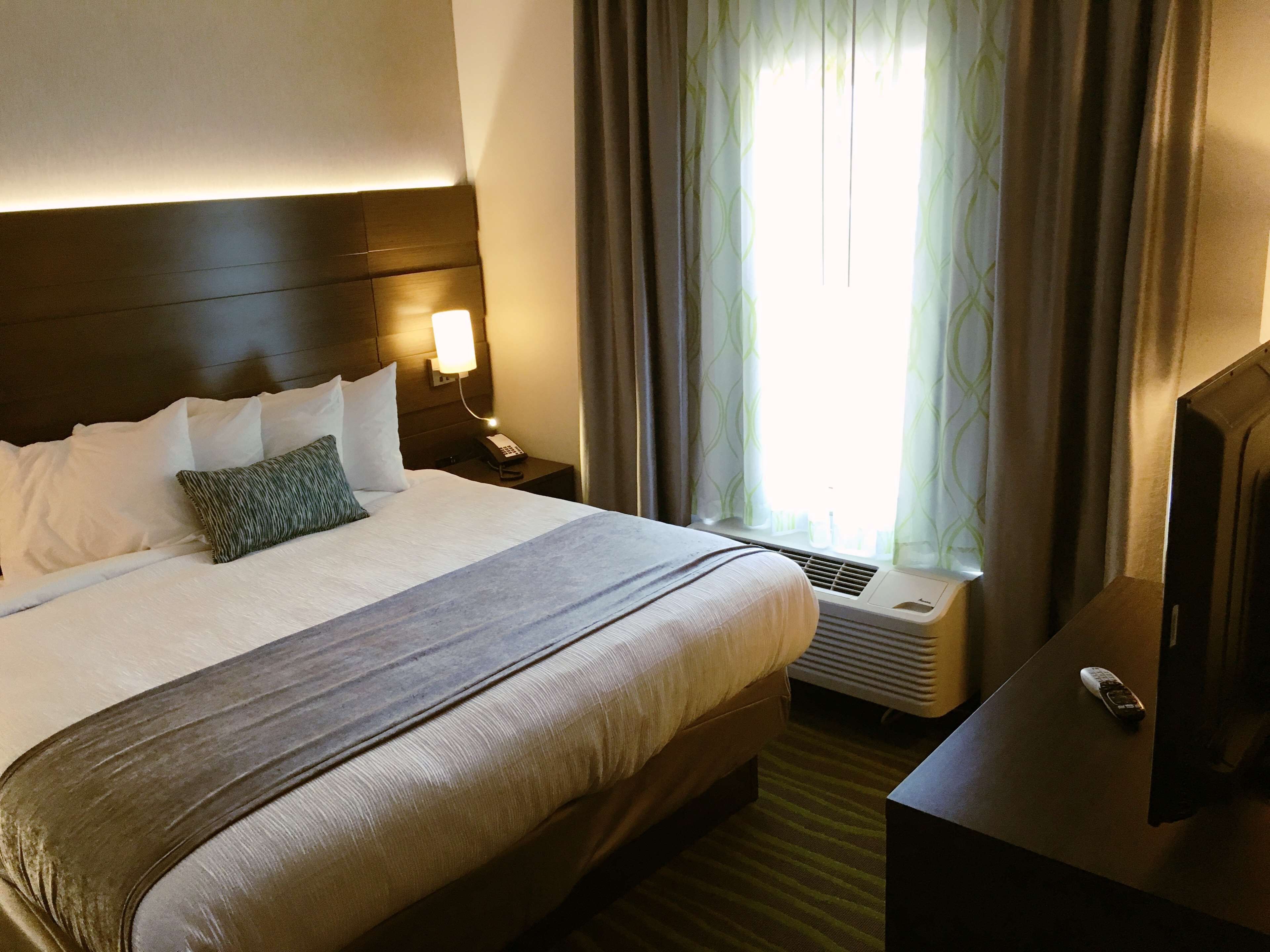 A hotel room with a large bed, a bedside lamp, a remote control on a nightstand, and a window with curtains.