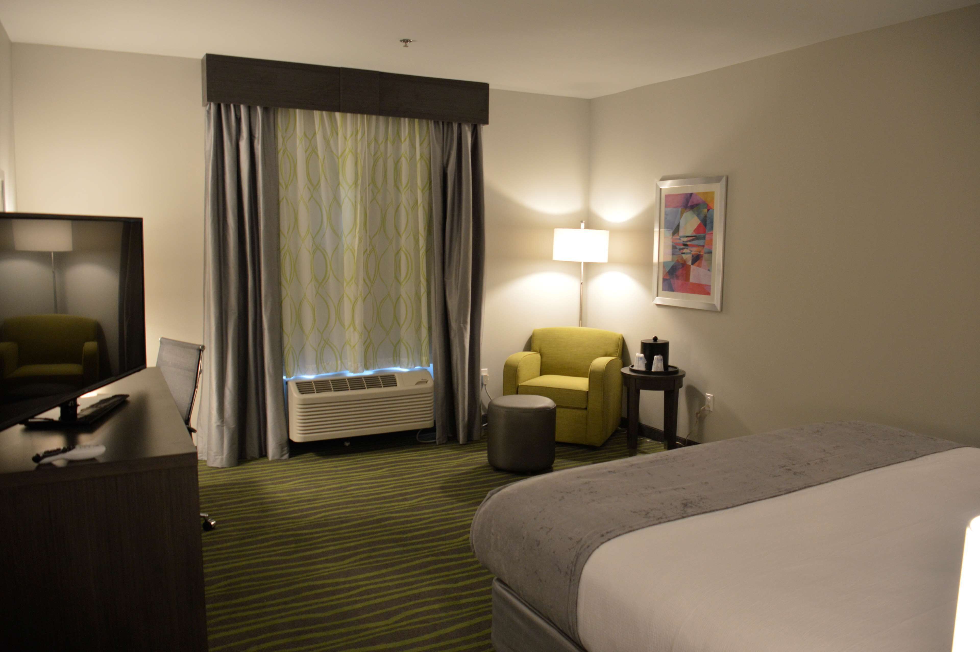 A hotel room with a bed, a desk with a computer, a chair, an air conditioner, and a lamp.