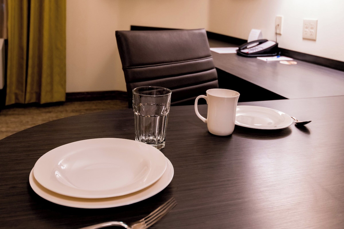 Habitación de hotel con una mesa de comedor preparada para dos personas, un televisor montado en la pared, un cuadro enmarcado y una lámpara.