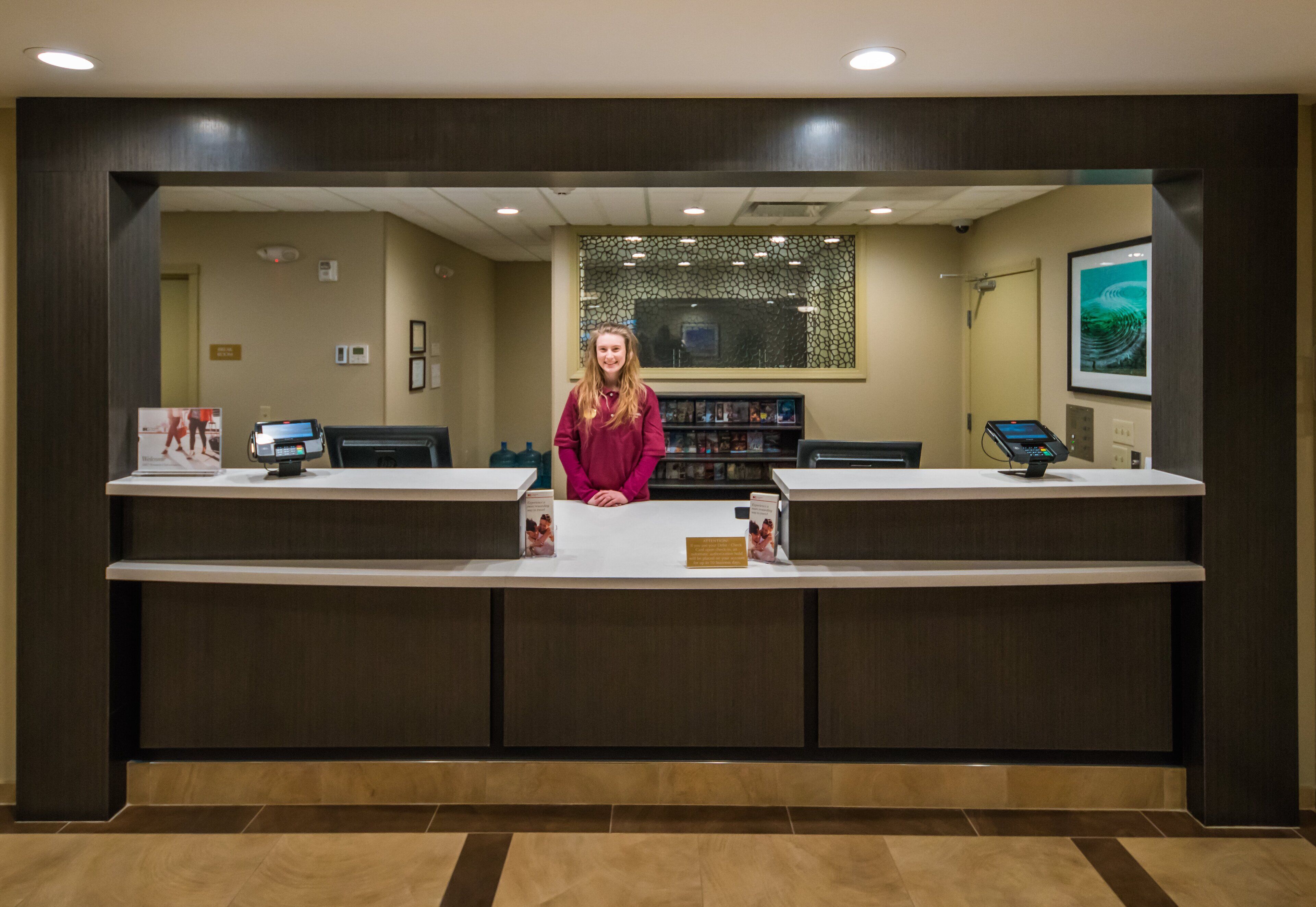 A reception desk with a woman standing behind it, a computer, and a framed picture on the wall.