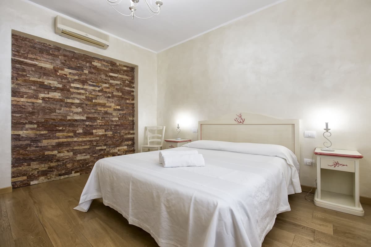 A hotel room with a wooden accent wall, a bed with white linens, a nightstand, a chair, and a wall-mounted air conditioner.