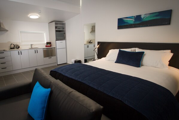 A modern hotel room with a large bed, a seating area with a blue pillow, a kitchenette, and a cityscape view. - Aurora On George (Dunedin)