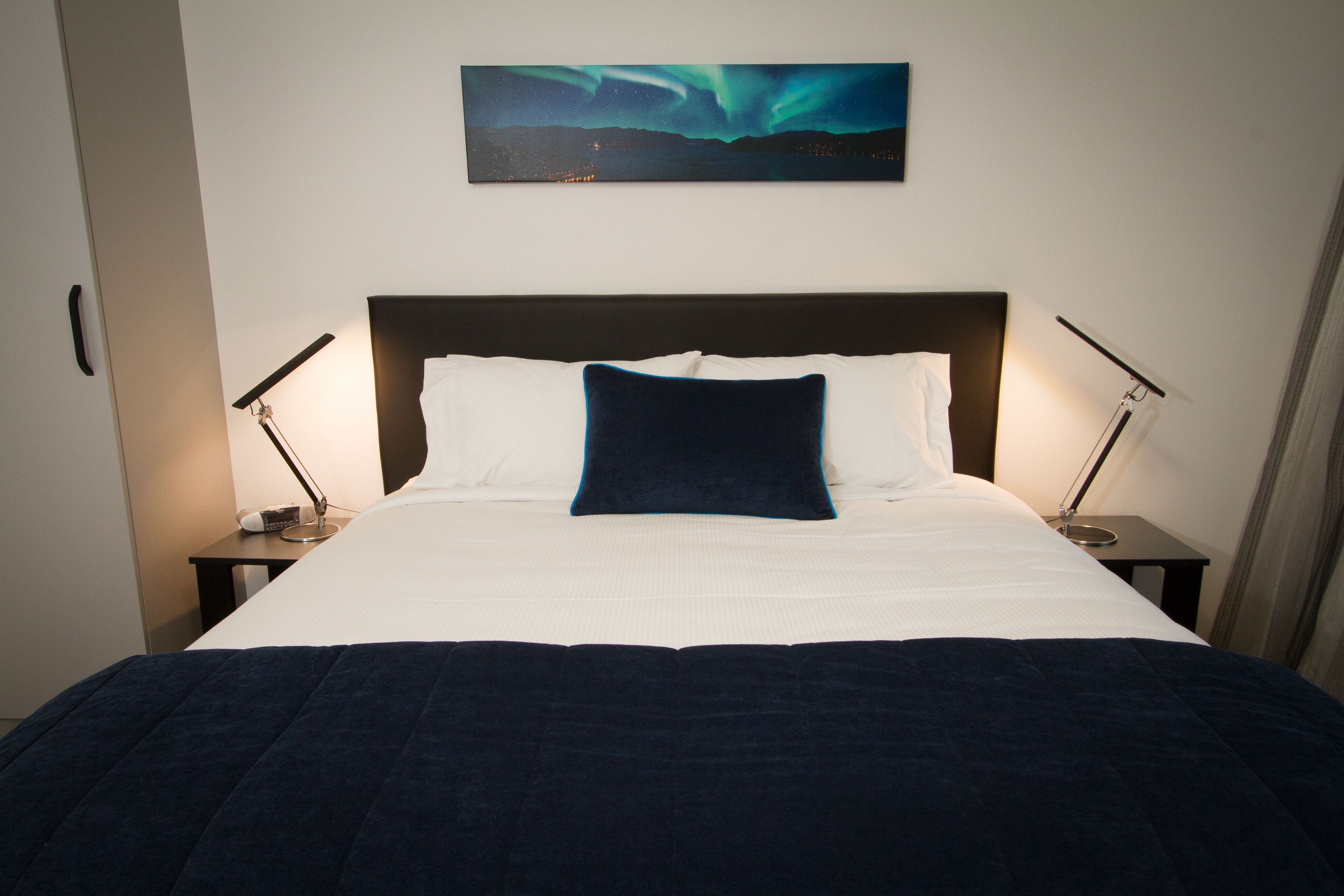 A neatly made bed with a dark blue blanket and pillows, two bedside lamps, and a painting of the Northern Lights above the bed.