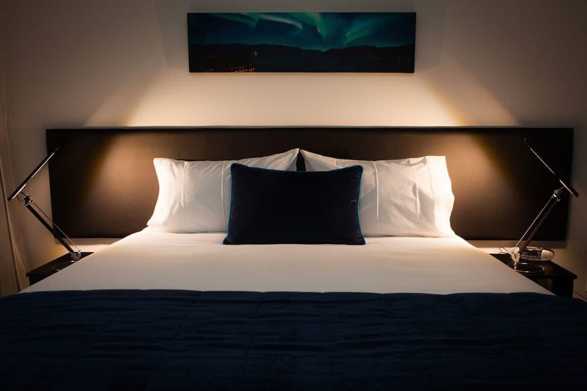 Una cama con ropa de cama blanca y un almohilar azul oscuro, flanqueada por dos mesitas de noche con lámparas, bajo un cuadro de la aurora boreal.