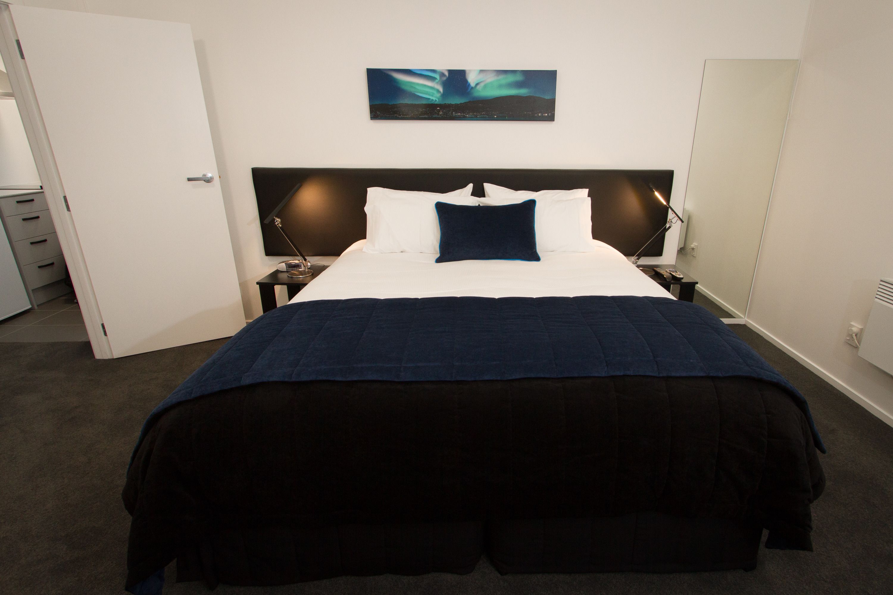 A hotel room with a large bed, a nightstand, a bedside lamp, a wall-mounted painting, and a door.