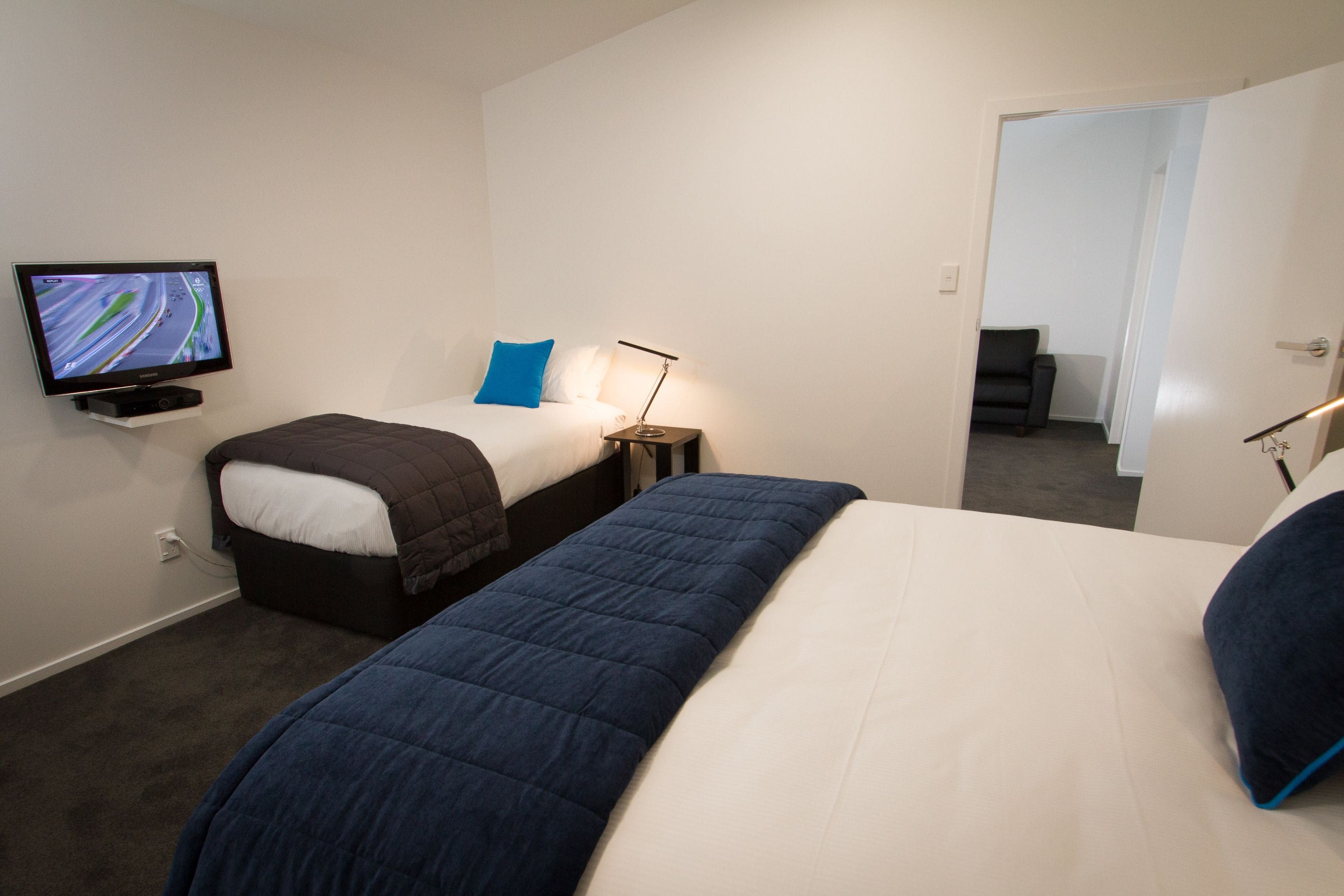 A hotel room with two beds, a TV on the wall, a desk with a lamp, and a chair in the corner.
