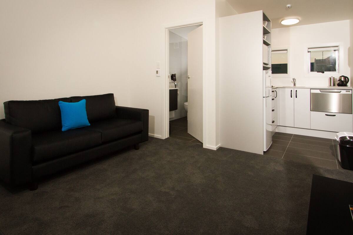 Una sala de estar moderna con un sofá negro y un cojín azul, una cocina con gabinetes blancos y electrodomésticos de acero inoxidable, y un piso cubierto de moqueta gris.