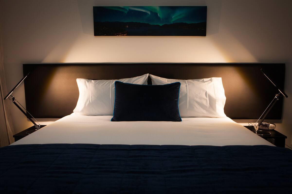 Una cama bien hecha con almohadas blancas y azul oscuro, cabecera oscura y un cuadro colgado en la pared de la aurora boreal.