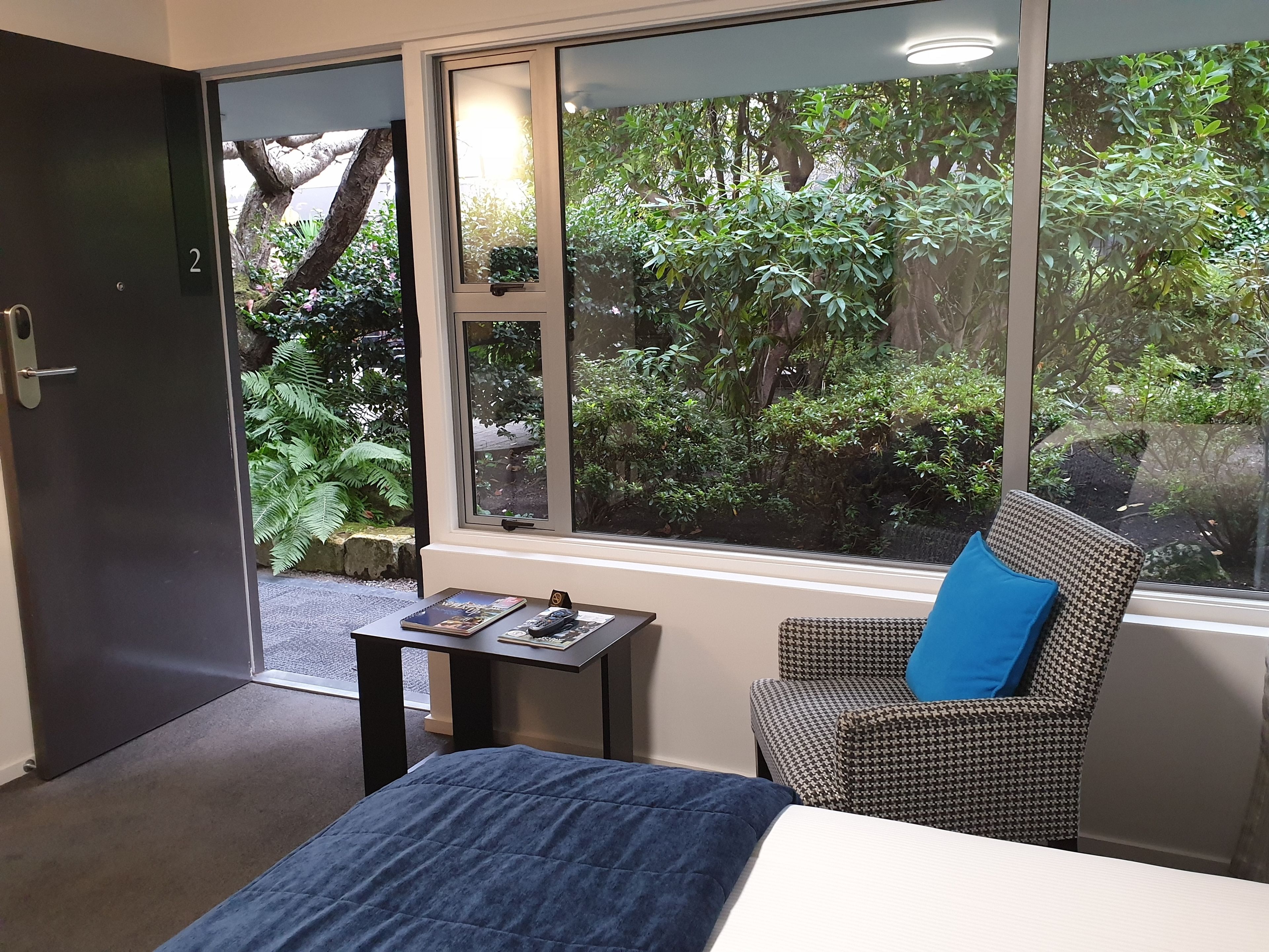 A room with a bed, a chair, a small table, and a window with a view of greenery.