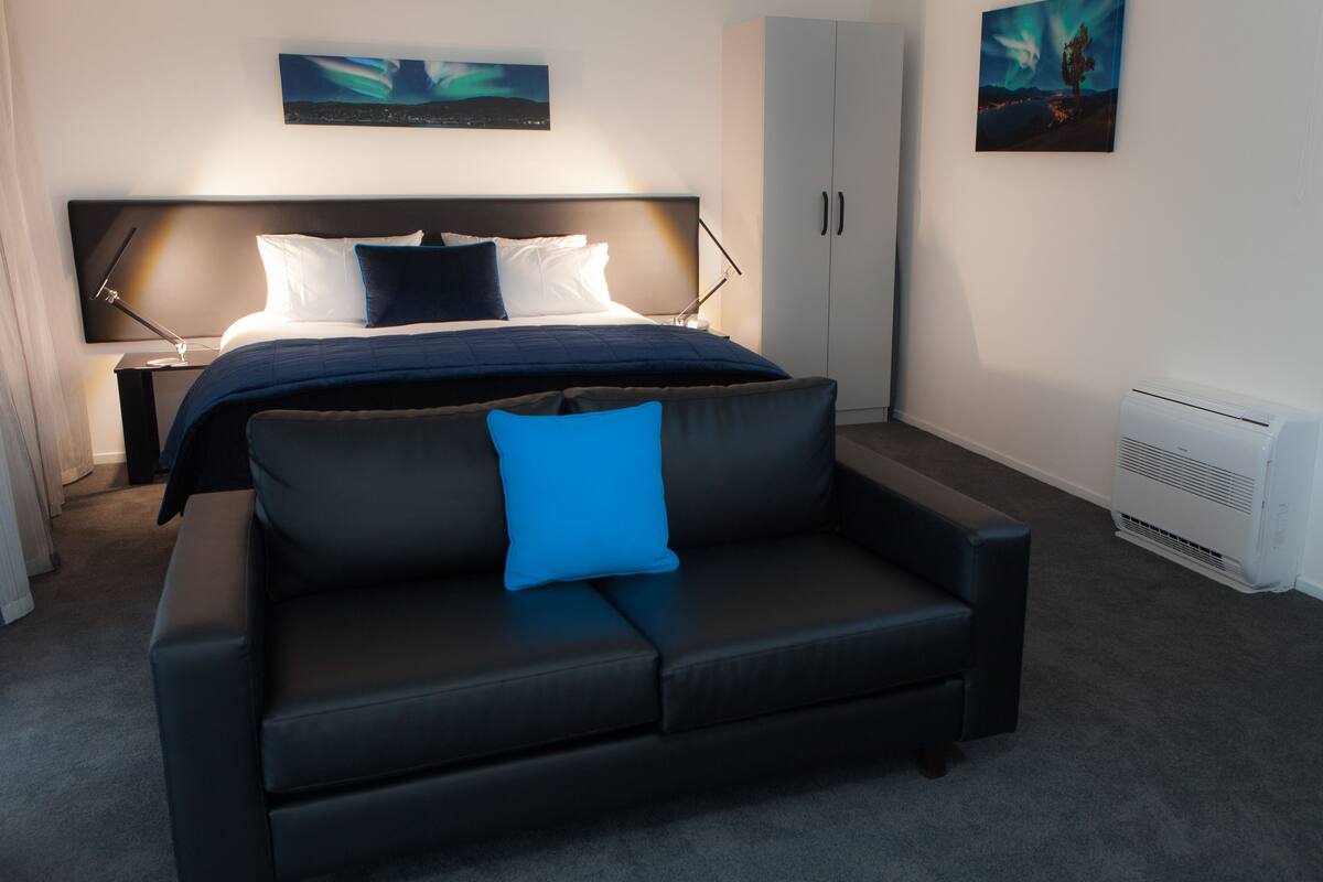 Una habitación de hotel moderna con una cama grande, un sofá de cuero oscuro y una mesita de noche con una lámpara.