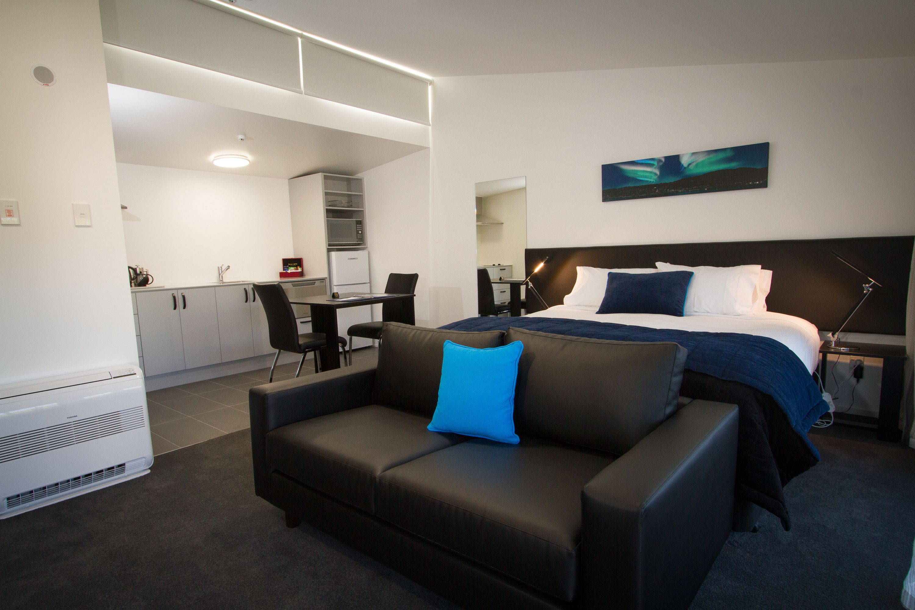 A modern hotel room with a bed, a sofa, a desk, and a kitchenette.